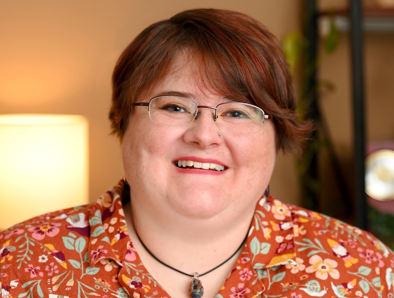 A person with short reddish-brown hair, glasses, and a nose piercing, smiling in a warmly lit room wearing a floral patterned shirt and a black choker necklace with a pendant.