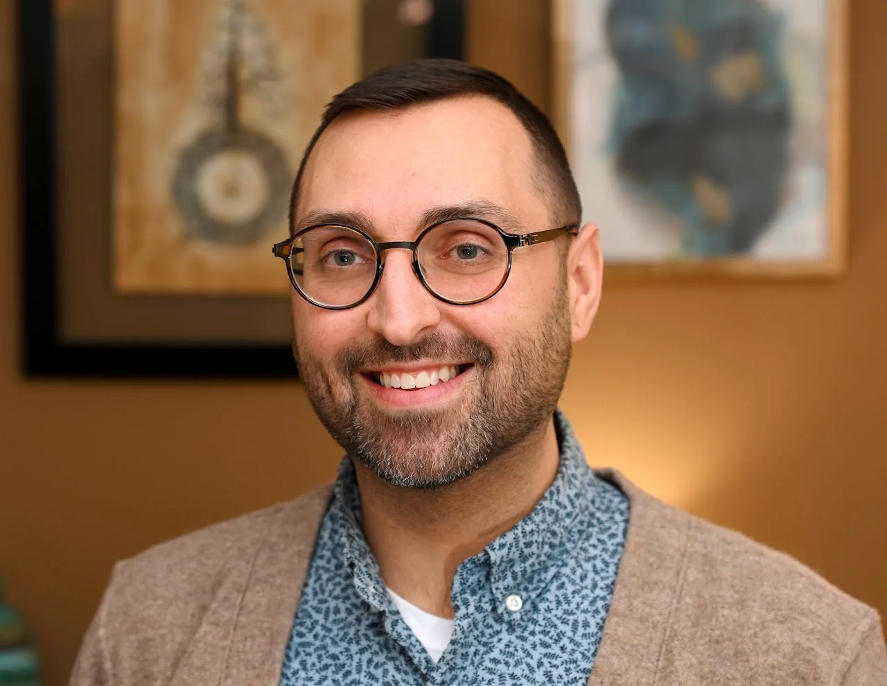 A smiling man with glasses, a beard, and short dark hair, wearing a patterned blue shirt and a beige sweater, in front of framed abstract art on a brown wall.