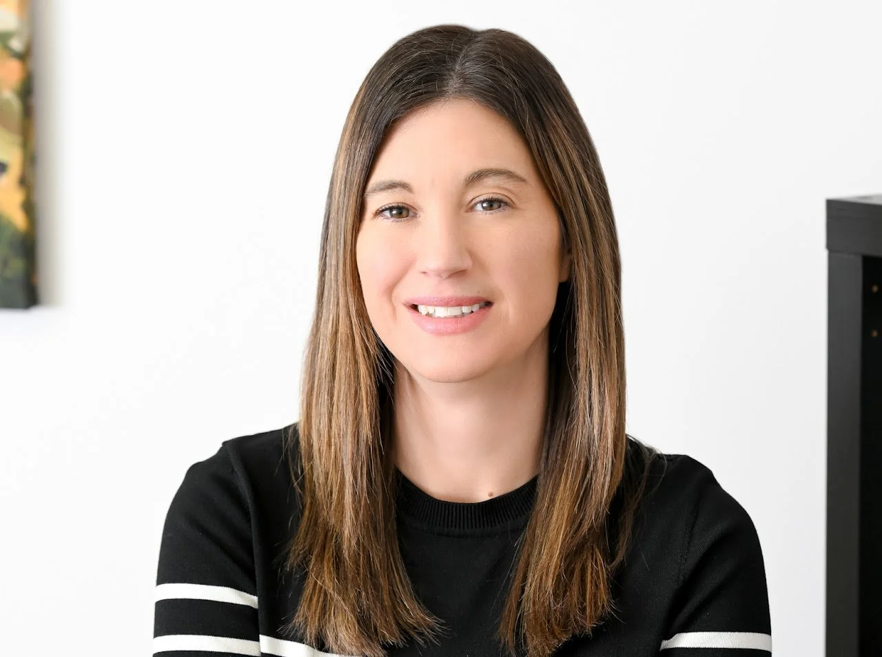 A woman with straight, shoulder-length brown hair, wearing a black shirt with white stripes on the sleeves, smiling at the camera in a bright, indoor setting.