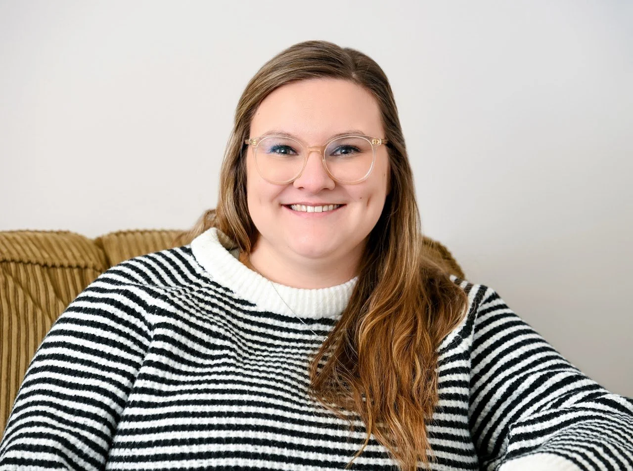 A woman with long, light brown hair and glasses smiling and sitting on a striped sweater on a beige couch.