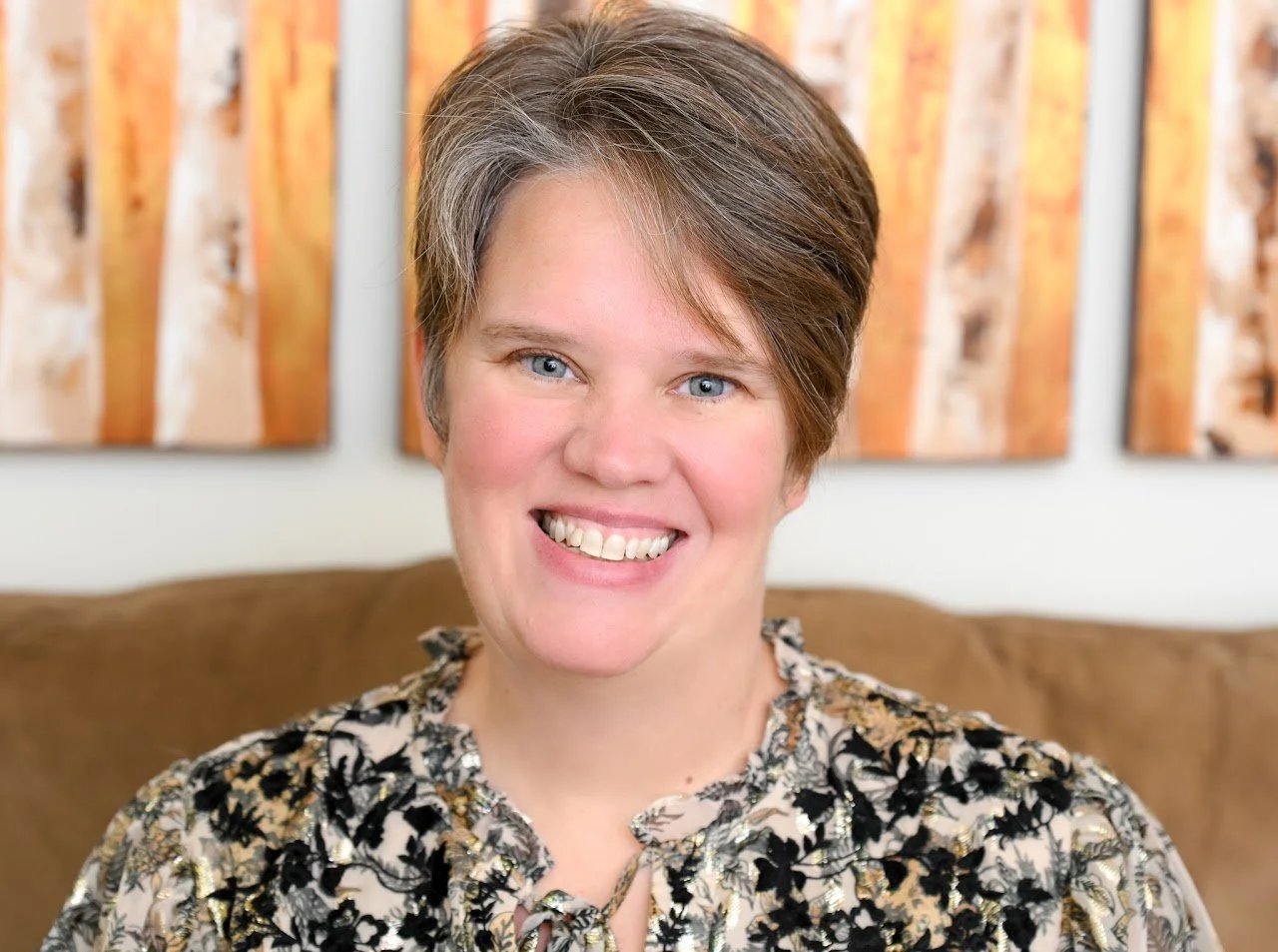 A woman with short brown hair and blue eyes, smiling, wearing a floral patterned blouse, sitting on a brown sofa with abstract orange and brown wall art in the background.