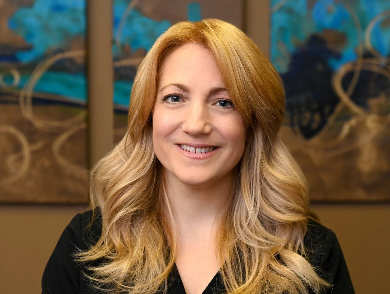 A woman with long blonde hair smiling at the camera, standing in front of abstract artwork with blue and brown tones.