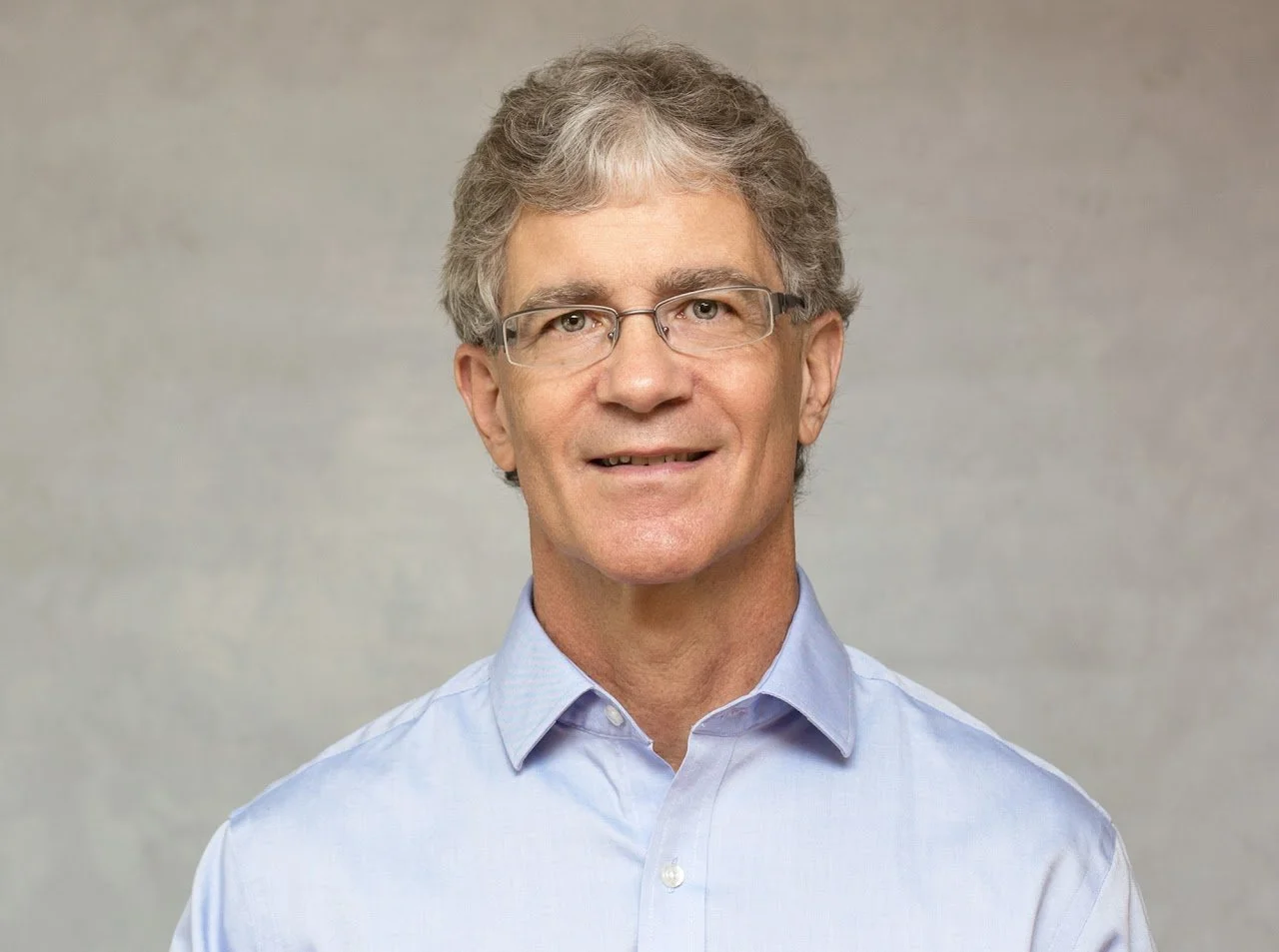 A middle-aged man with curly gray hair and glasses, wearing a light blue collared shirt, smiling at the camera against a plain gray background.