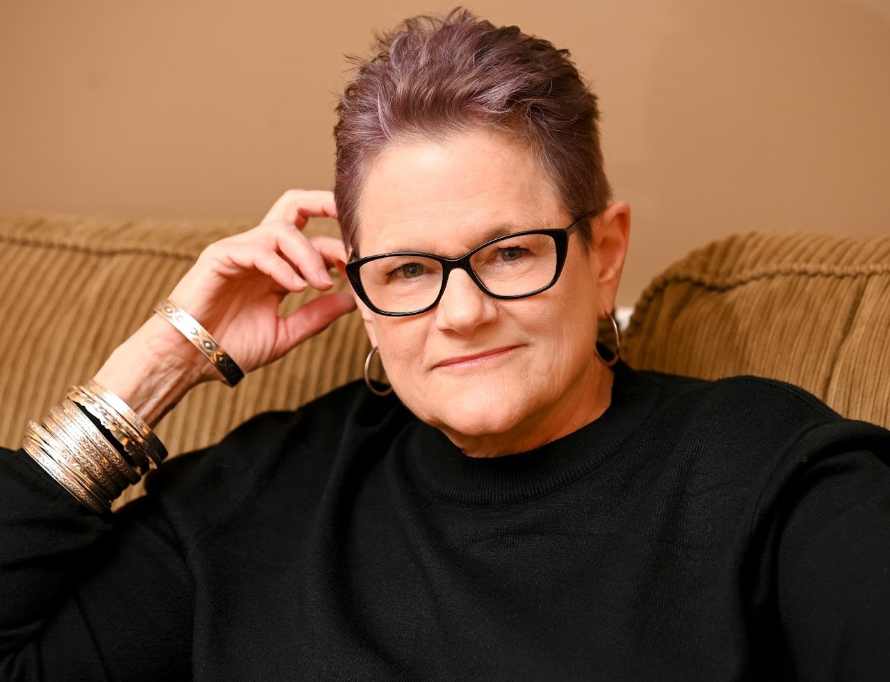 A mature woman with short gray hair, black glasses, and a black top, sitting on a tan couch with her hand near her ear, wearing jewelry including earrings and multiple bracelets.