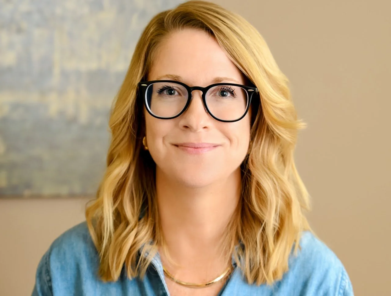 A woman with blonde, wavy hair wearing black-rimmed glasses, a denim shirt, and a gold necklace, smiling at the camera.