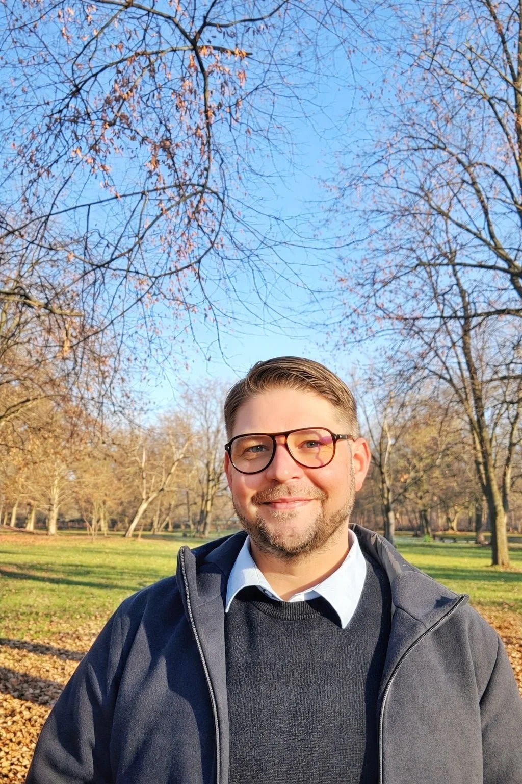 A man with glasses smiling outdoors in a park with trees and fallen leaves, clear blue sky