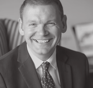 Black and white photo of a smiling man in a suit with a patterned tie, indoors.