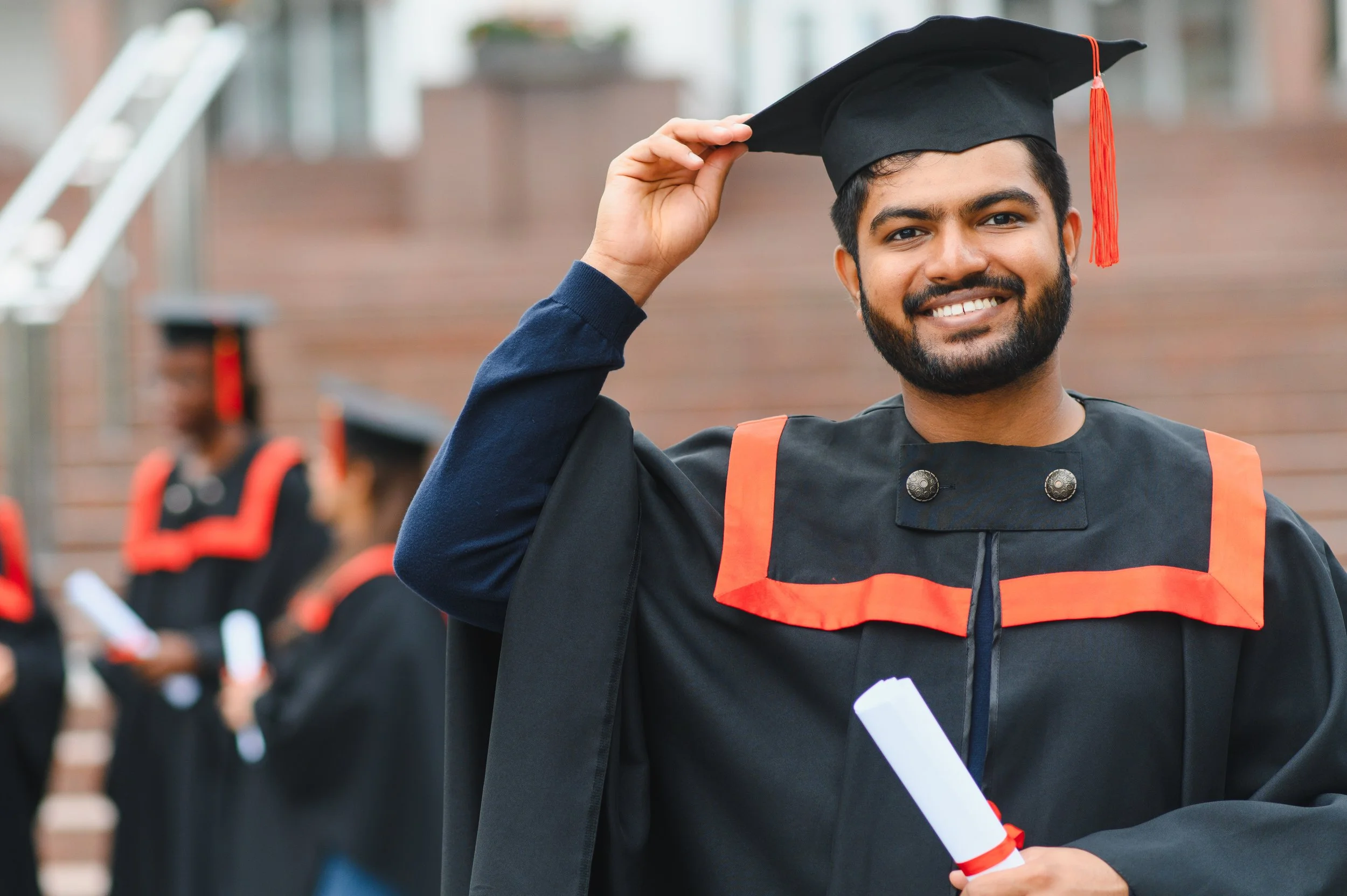 A young man in graduation robes and mortarboard smiling and holding his graduation certificate, standing outdoors with other graduates in the background.