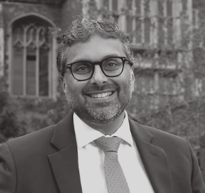 A smiling man with glasses and a beard, wearing a suit and tie, standing outdoors in front of a building with arched windows.