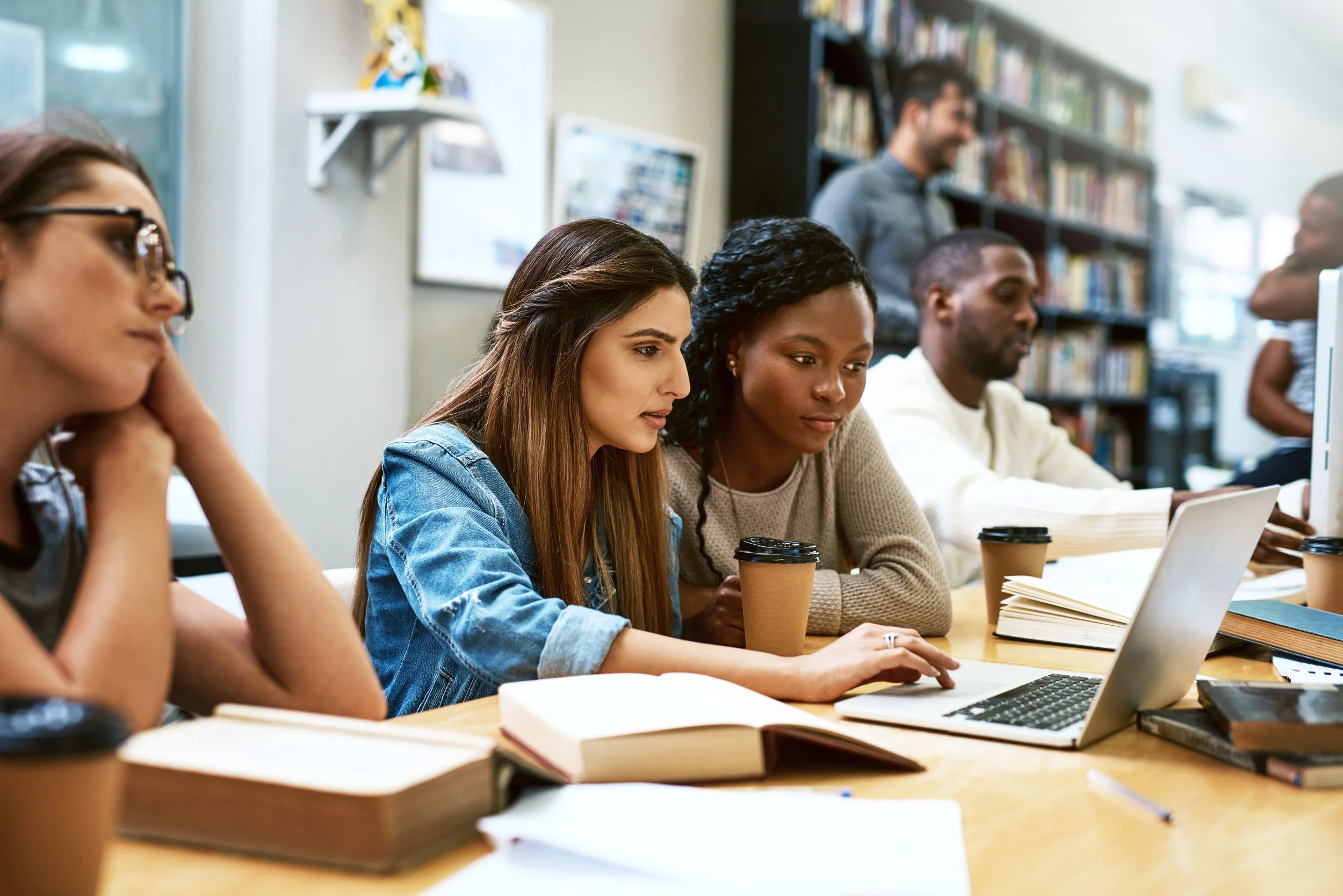 A diverse group of people sitting at a table in a library or study room, focused on laptops and surrounded by books, with some holding coffee cups.