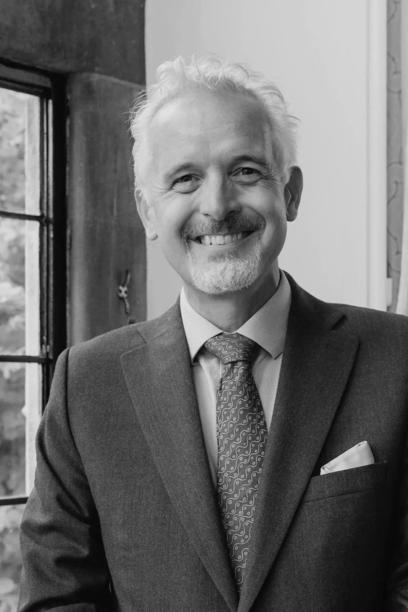 A smiling man with white hair and a beard, wearing a suit, tie, and pocket square, standing indoors near a window and curtain.