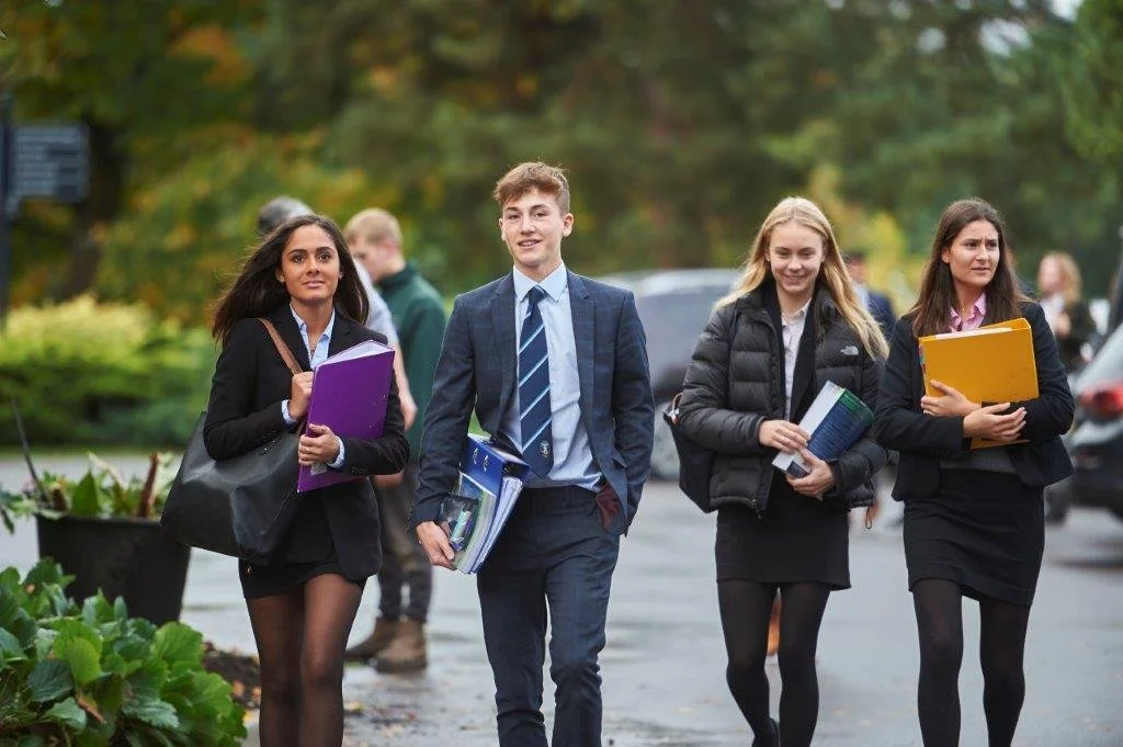 Four students walking outdoors on a school day, carrying folders and books, with autumn trees in the background.