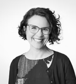 Black and white portrait of a woman with curly hair and glasses, smiling, wearing a patterned blouse and a cardigan.