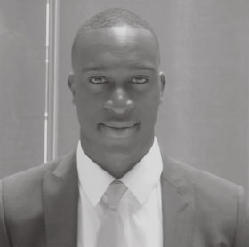 A black-and-white portrait of a man in a suit and tie, looking at the camera with a neutral expression.