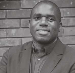 Black and white portrait of a smiling man wearing a suit jacket and a shirt, standing in front of a brick wall.