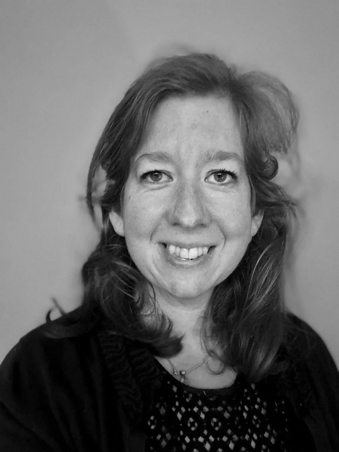 Black and white portrait of a woman with shoulder-length wavy hair, smiling, wearing a patterned top and a necklace, against a plain background.