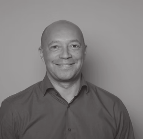 Black and white portrait of a smiling man with a shaved head, wearing a collared shirt, against a plain background.