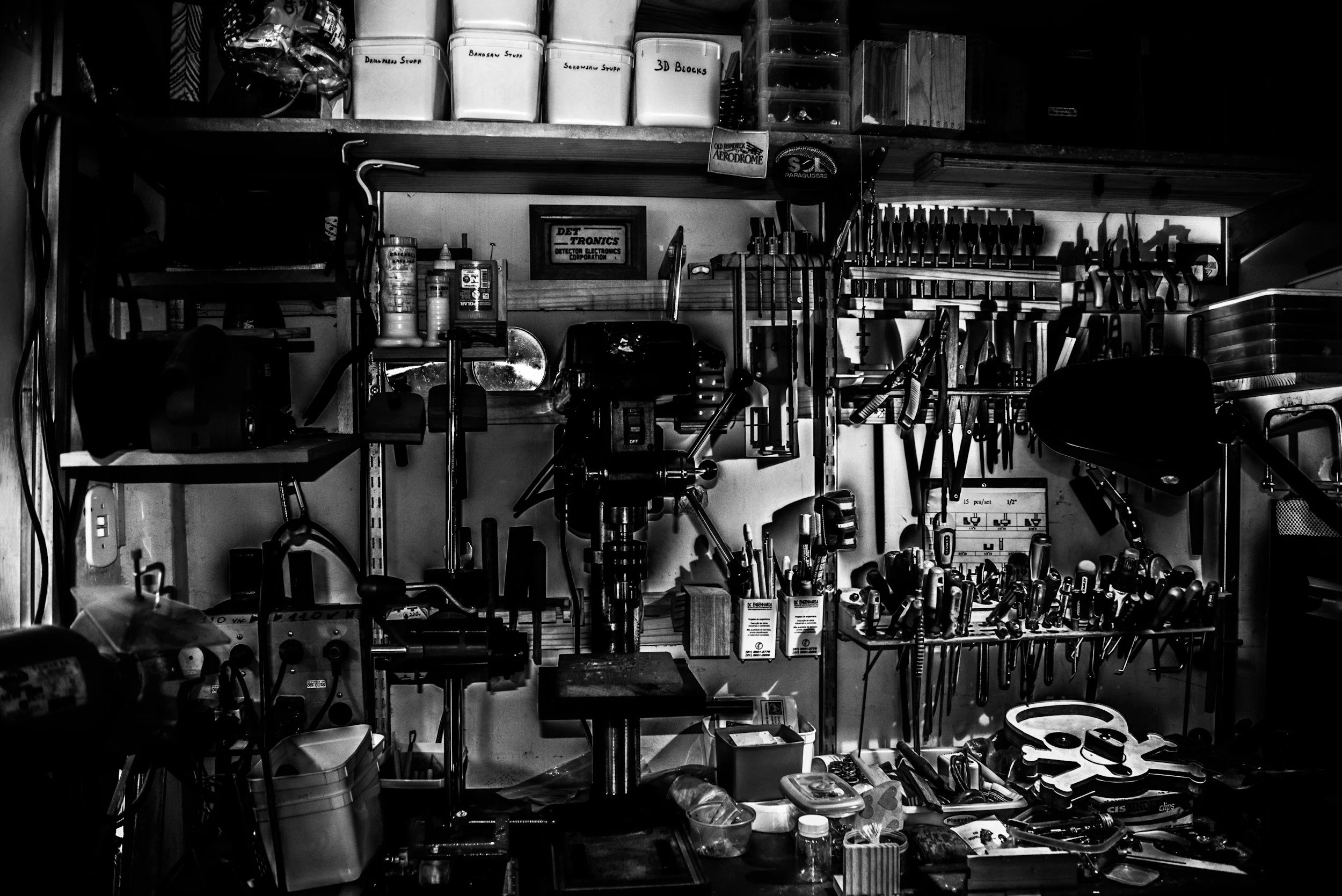 A black-and-white photo of a cluttered workshop or garage shelf filled with tools, storage containers, and miscellaneous items.