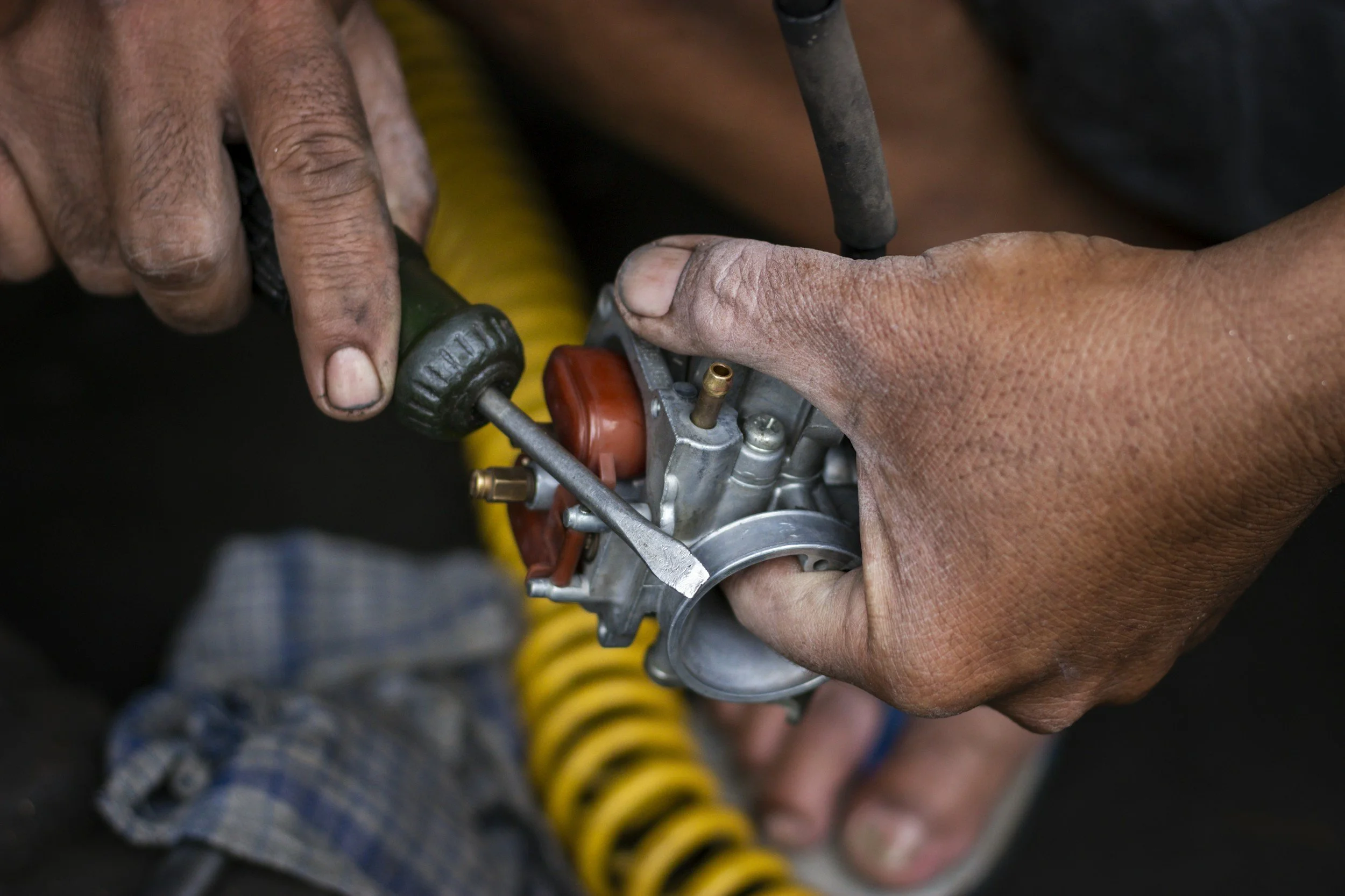 Close-up of a person's hands working on a small mechanical or engine part, with some tools and a yellow and black striped object in the background.
