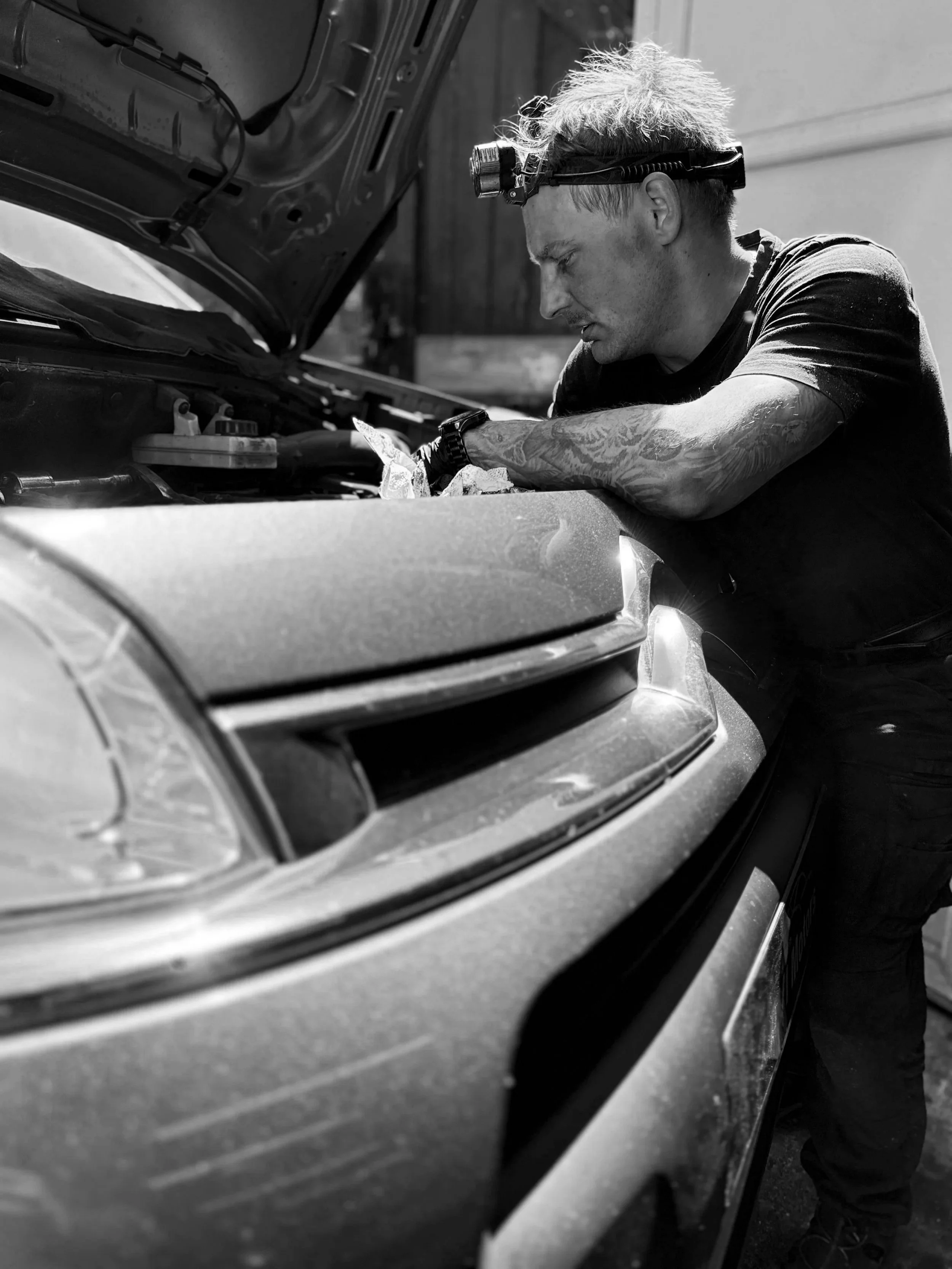 A man with tattoos and a headlamp working underneath the open hood of a car.