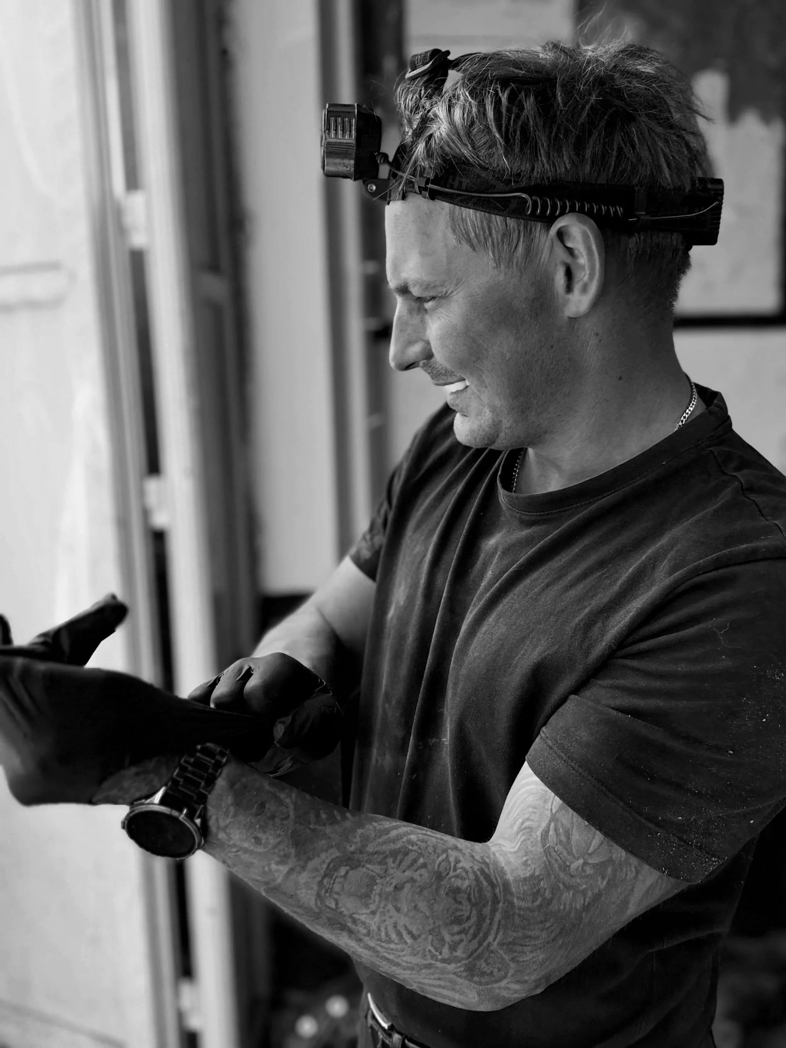 A man with tattoos on his arms looking at his wristwatch, wearing a black shirt and a headlamp, with a background of a workspace.