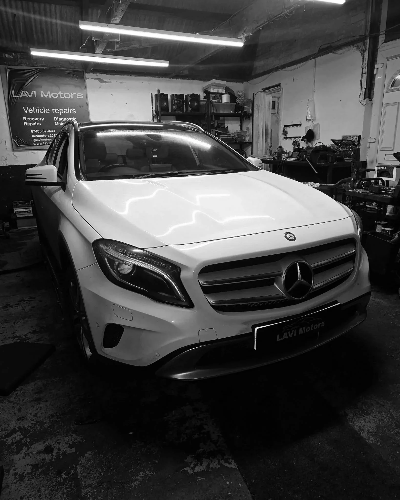 A white Mercedes-Benz car parked inside a garage with tools and shelves in the background. A banner on the wall advertises vehicle repair services.