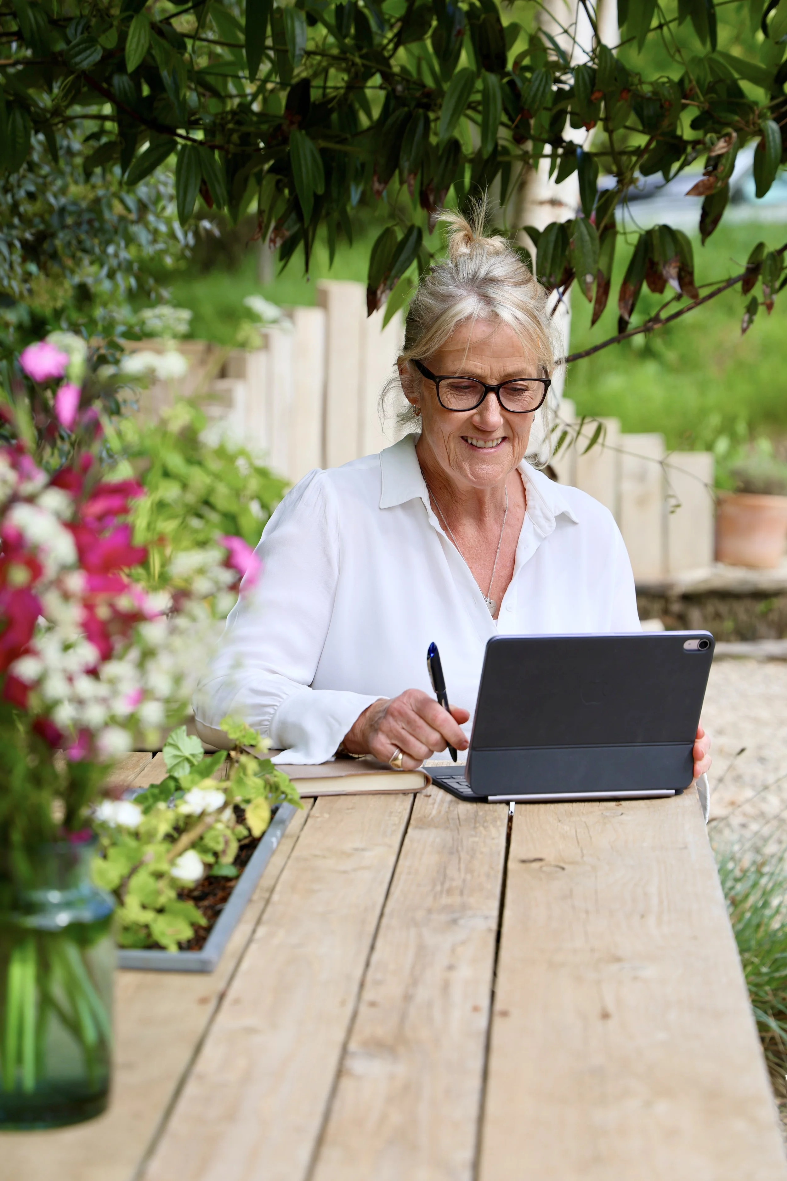 A woman with glasses sitting at a wooden outdoor table in a garden, using a tablet with a stylus, surrounded by colourful flowers and green foliage.