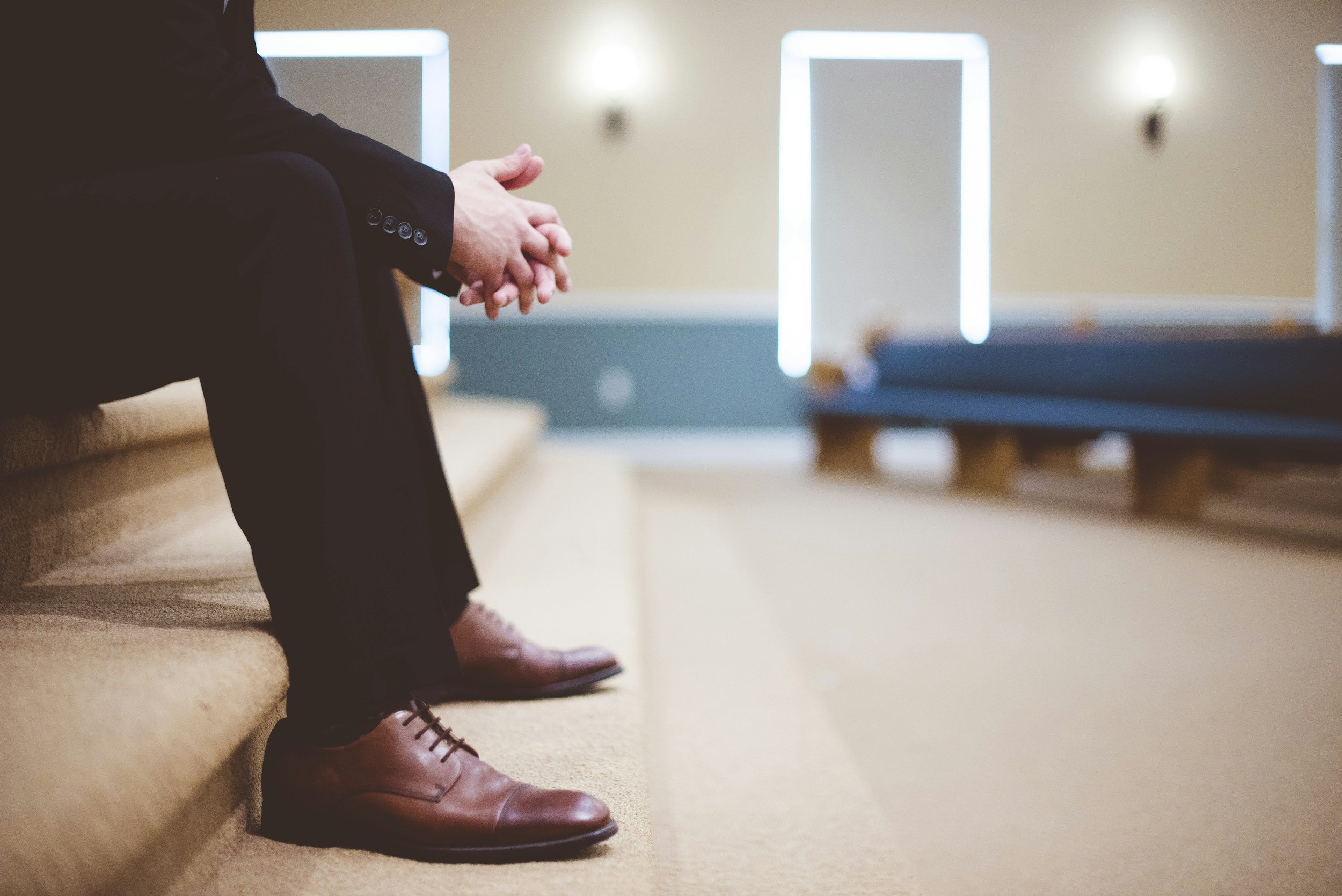 Someone sitting on a beige couch with hands clasped, dressed in black pants and brown shoes, in a room with wall-mounted lights and a blue bench.
