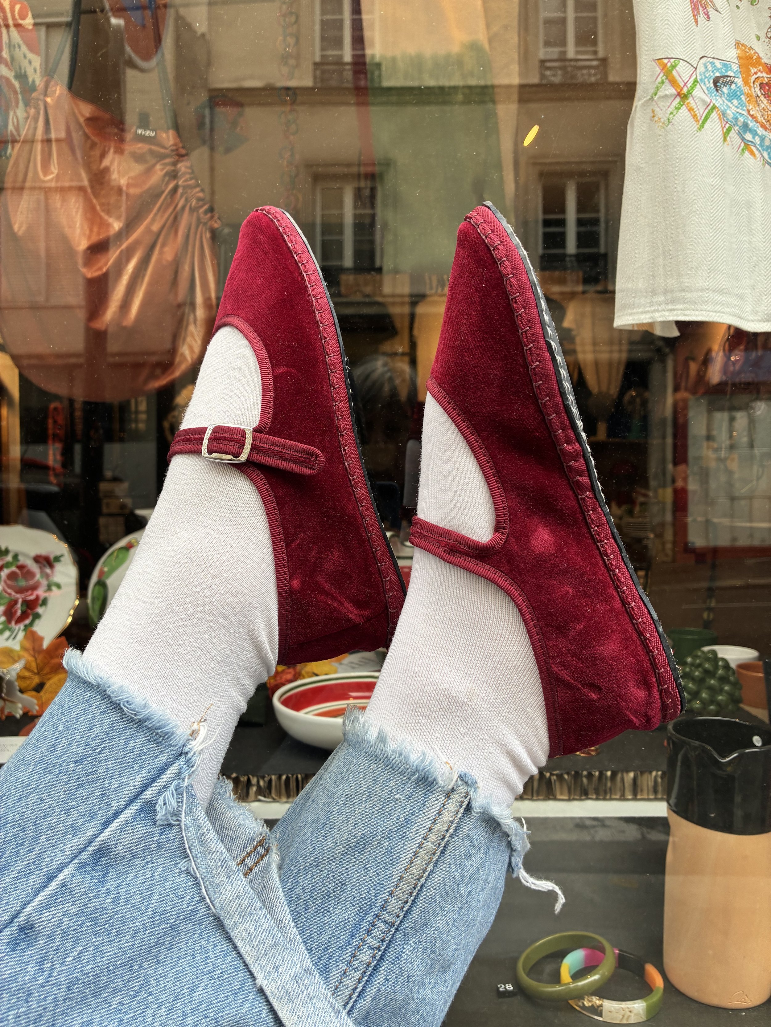 Une personne portant des chaussures rouges en velours et des jeans déchirés regarde par une fenêtre d'une boutique, avec divers objets décoratifs et céramiques visibles à l'intérieur.
