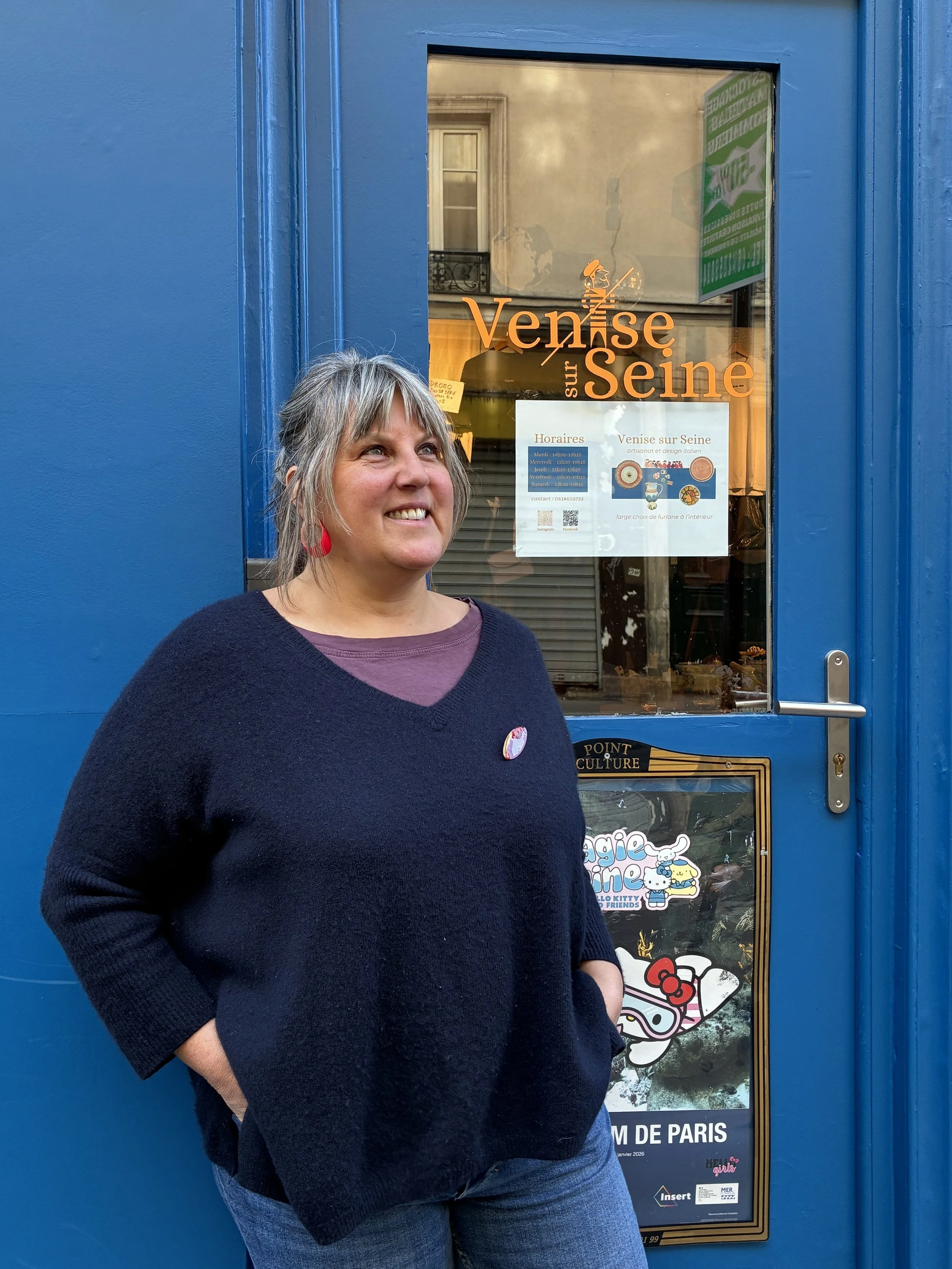 Une femme souriante avec des cheveux gris, portant un pull noir et des boucles d'oreilles rouges, se tient devant une porte bleue avec des affiches d'événements culturels et des personnages de dessins animés.