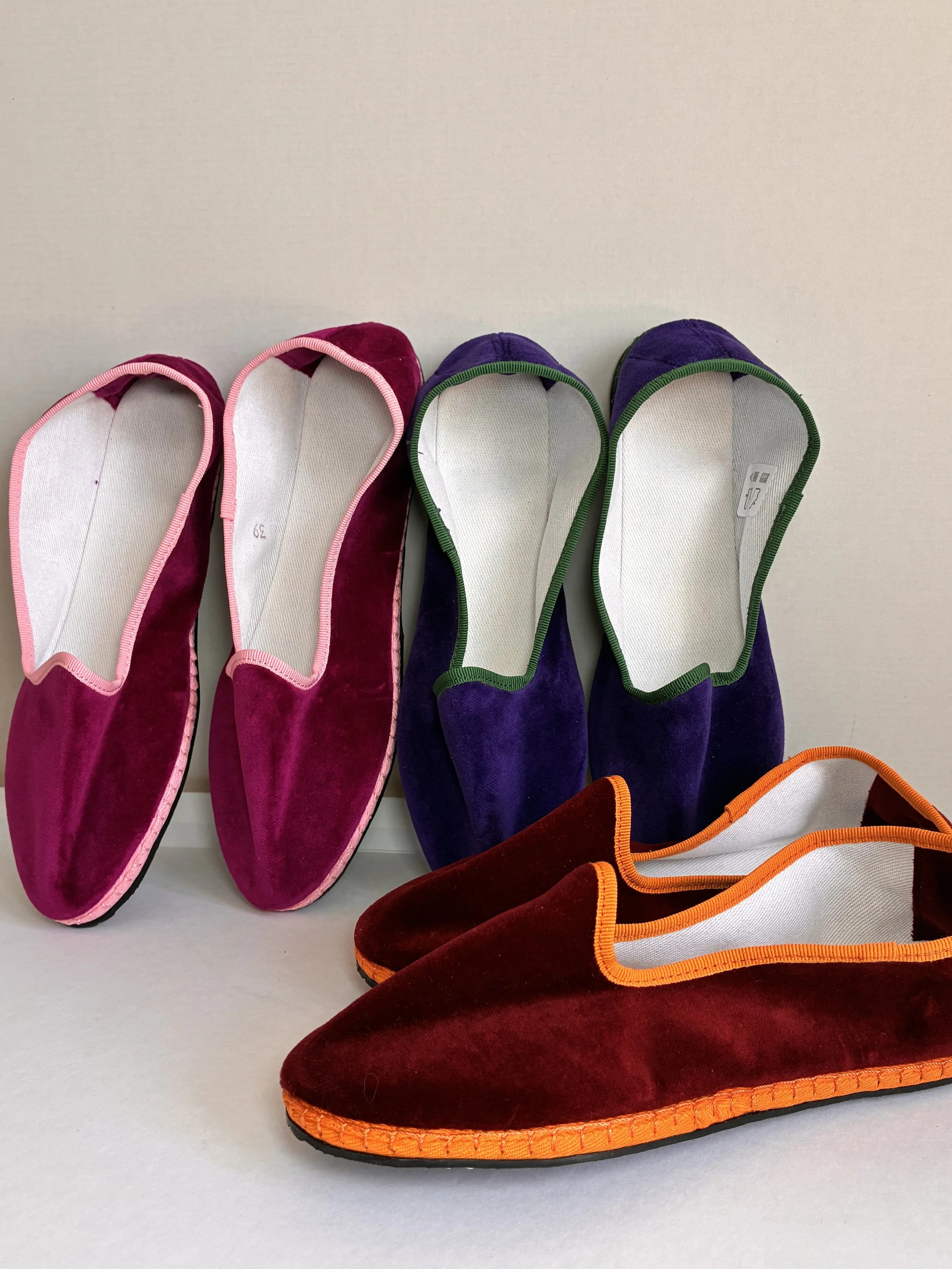 Chaussures plates en velours de couleur rose, violette et bordeaux avec double bordure colorée, posées sur une surface blanche.