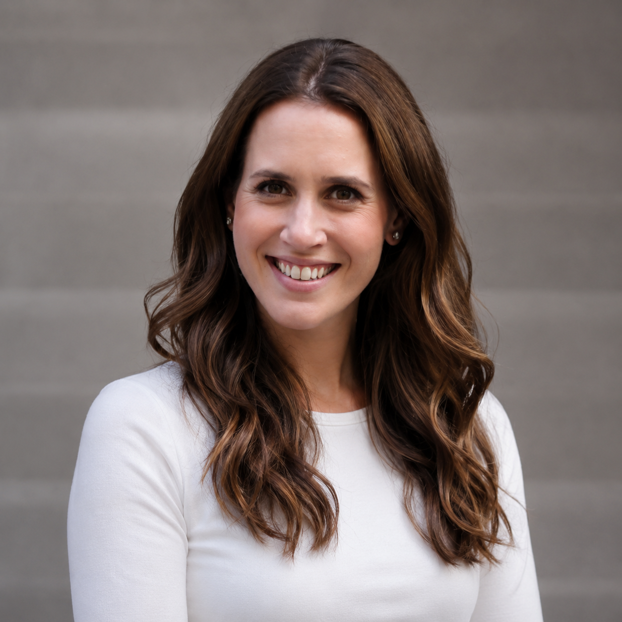 A woman with long, wavy brown hair smiling, wearing a white top, standing against a grey background.