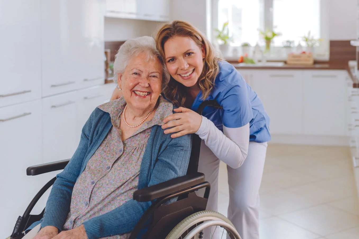 A young female caregiver in blue scrubs smiling with an elderly woman in a wheelchair in a bright kitchen.