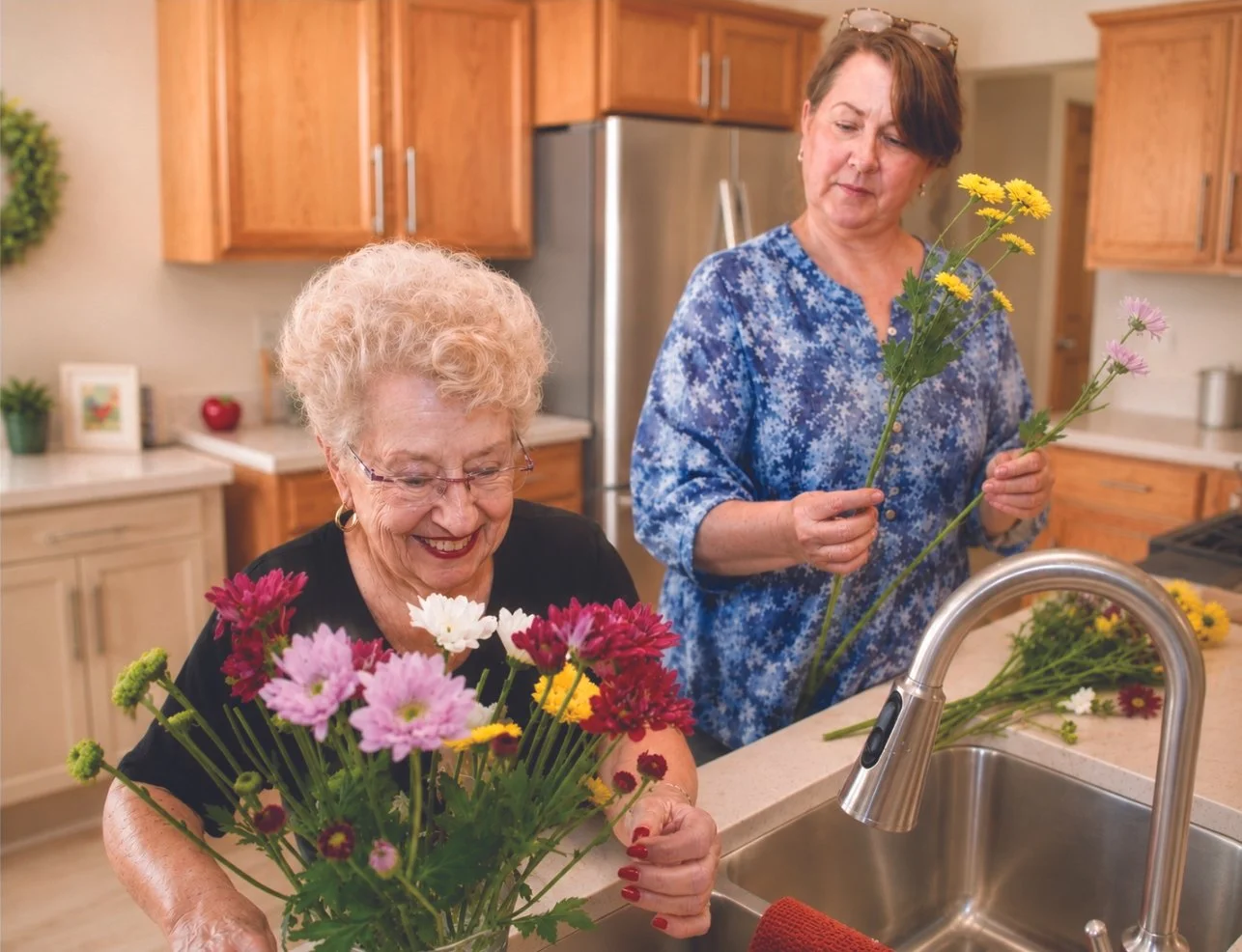 Two women in a kitchen arranging and admiring colorful flowers in a vase