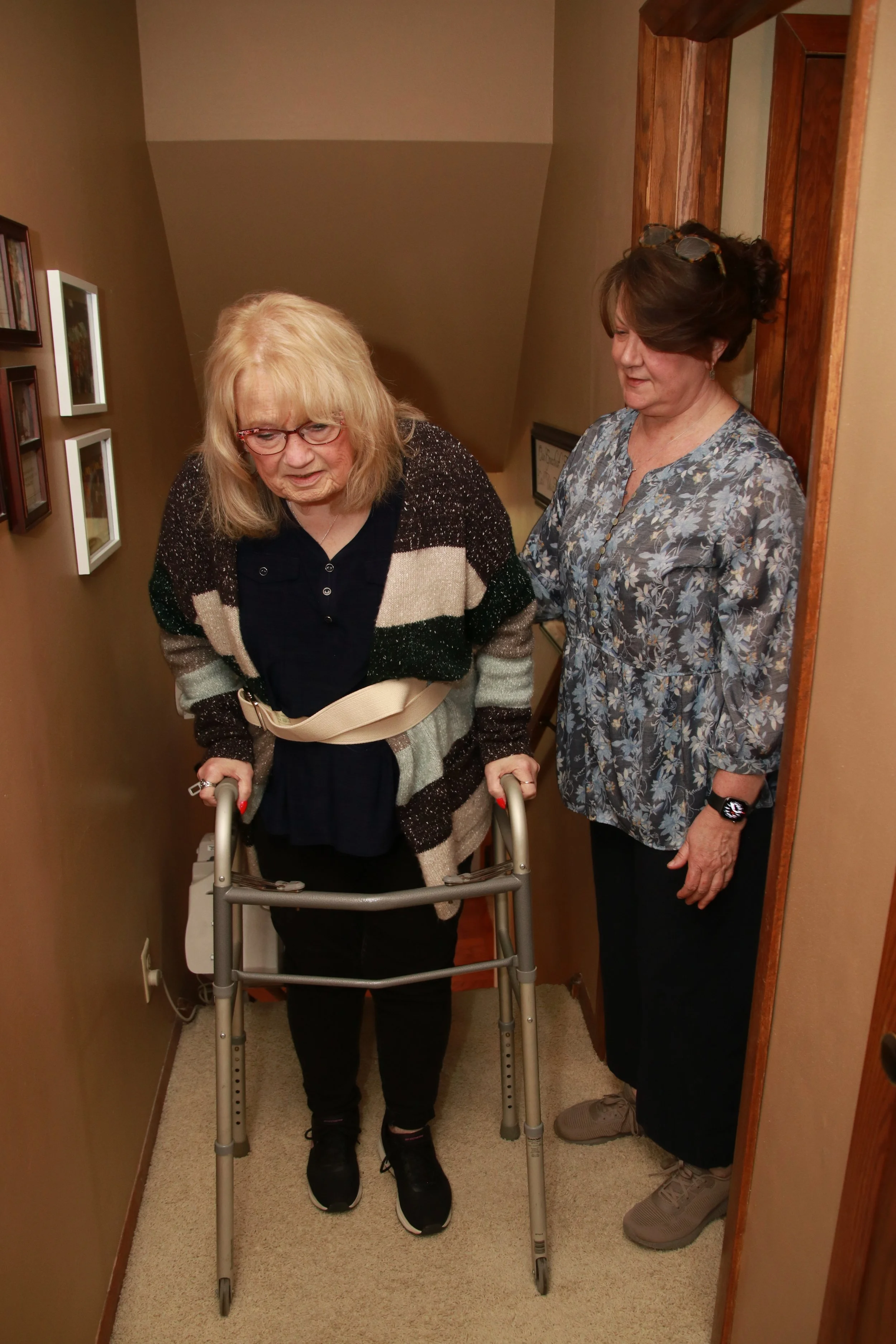 An elderly woman with blonde hair, wearing glasses, a black shirt, and a striped cardigan, using a walker down a narrow hallway. A middle-aged woman with dark hair, wearing a patterned blouse and glasses on her head, stands to her right, providing support.