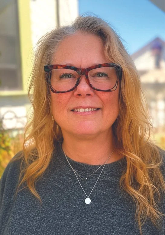 A smiling woman with wavy blonde hair and glasses standing outdoors with a house and blue sky in the background.