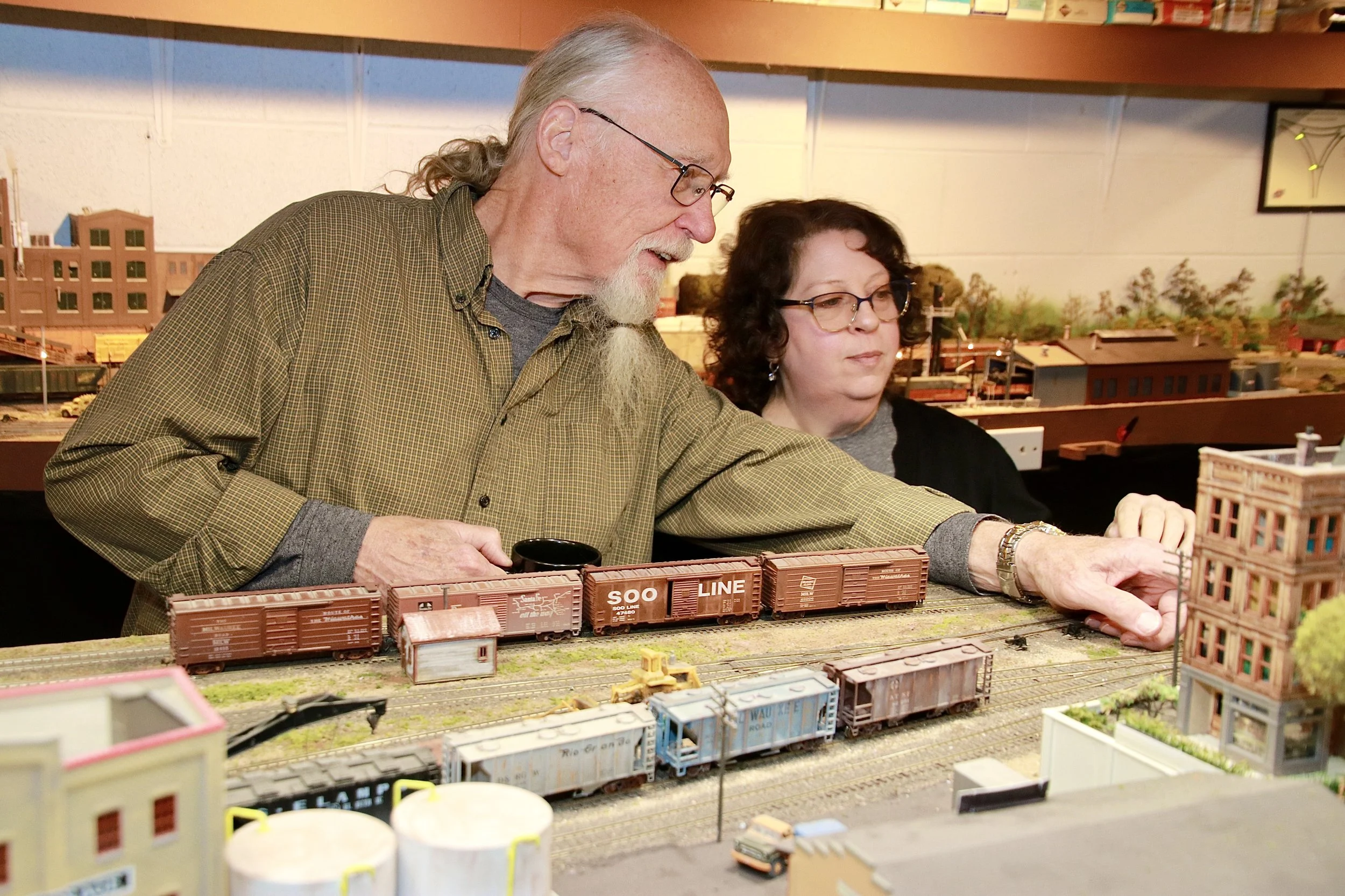 Two older adults, a man with long gray hair and beard wearing glasses and a man with curly hair and glasses, are working together on a detailed model train set, with various train cars and miniature buildings.