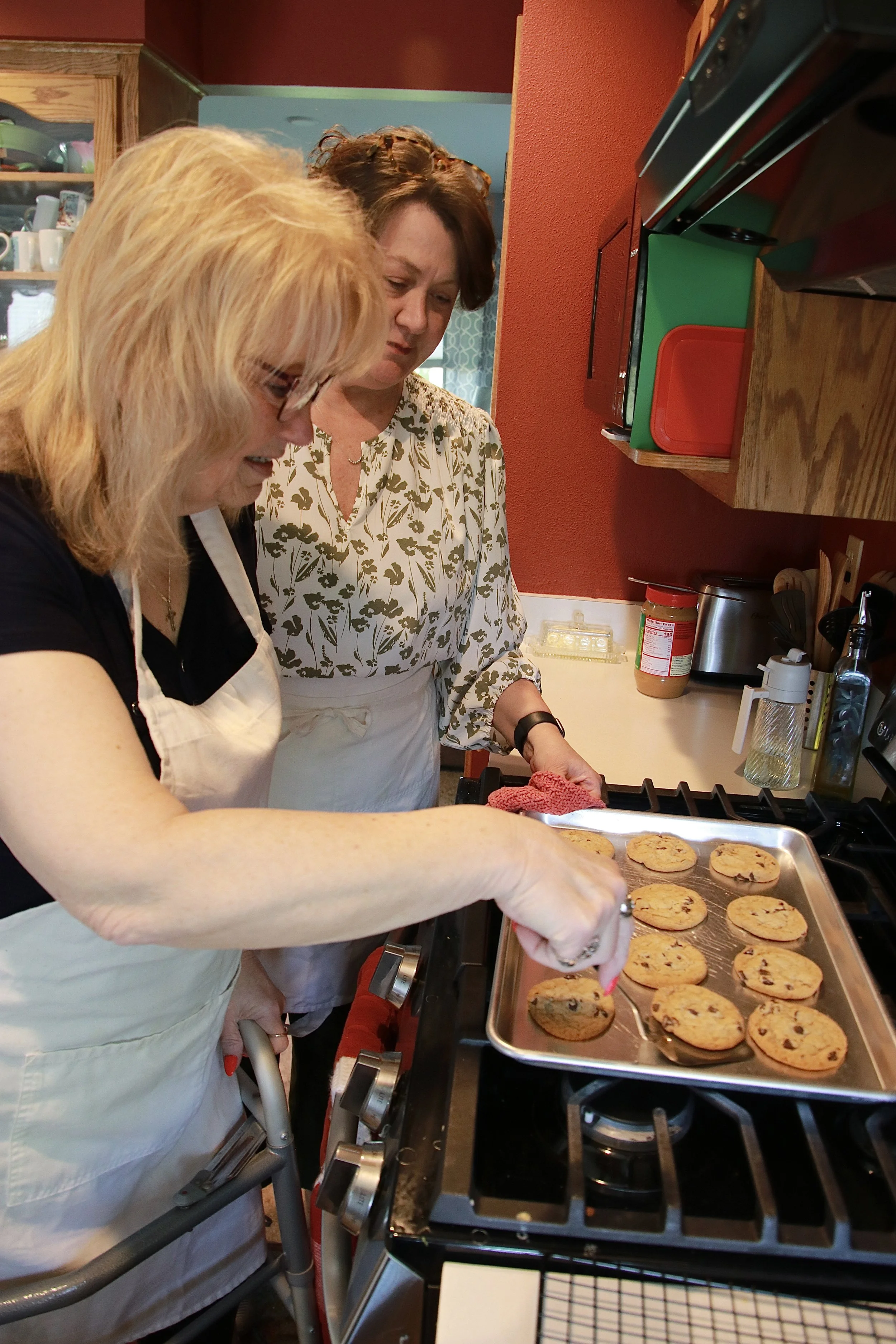 Two women baking cookies together in a kitchen. One woman, with blonde hair, is reaching into a rack of cookies on a baking sheet. The other woman, with short reddish-brown hair, is standing beside her. The kitchen has wooden cabinets and a red wall, with various kitchen items around.