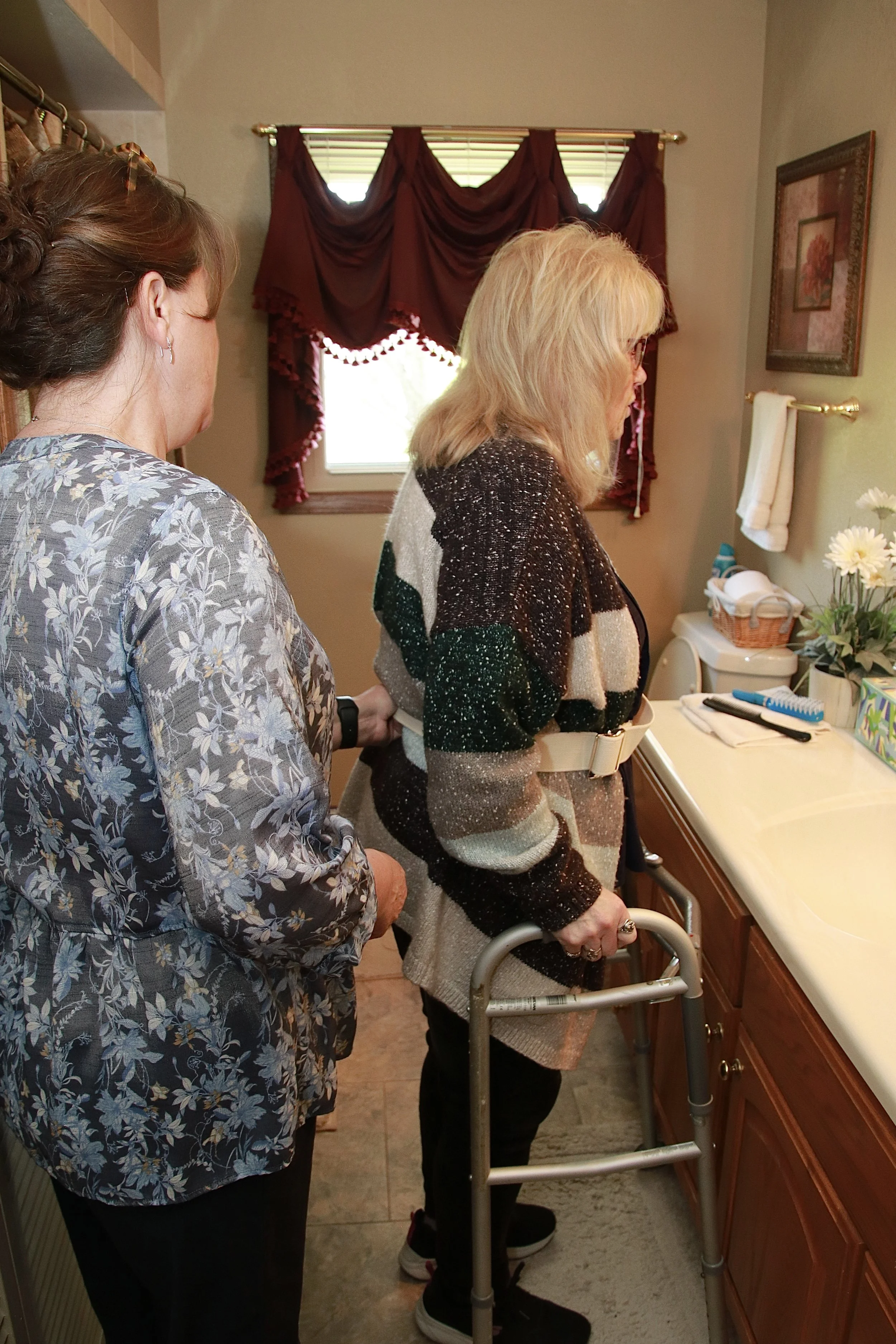 A woman with blonde hair using a walker stands at a bathroom sink, with another woman with brown hair standing beside her, both in a bathroom with a window and red curtains, and a vanity with flowers and toiletries.