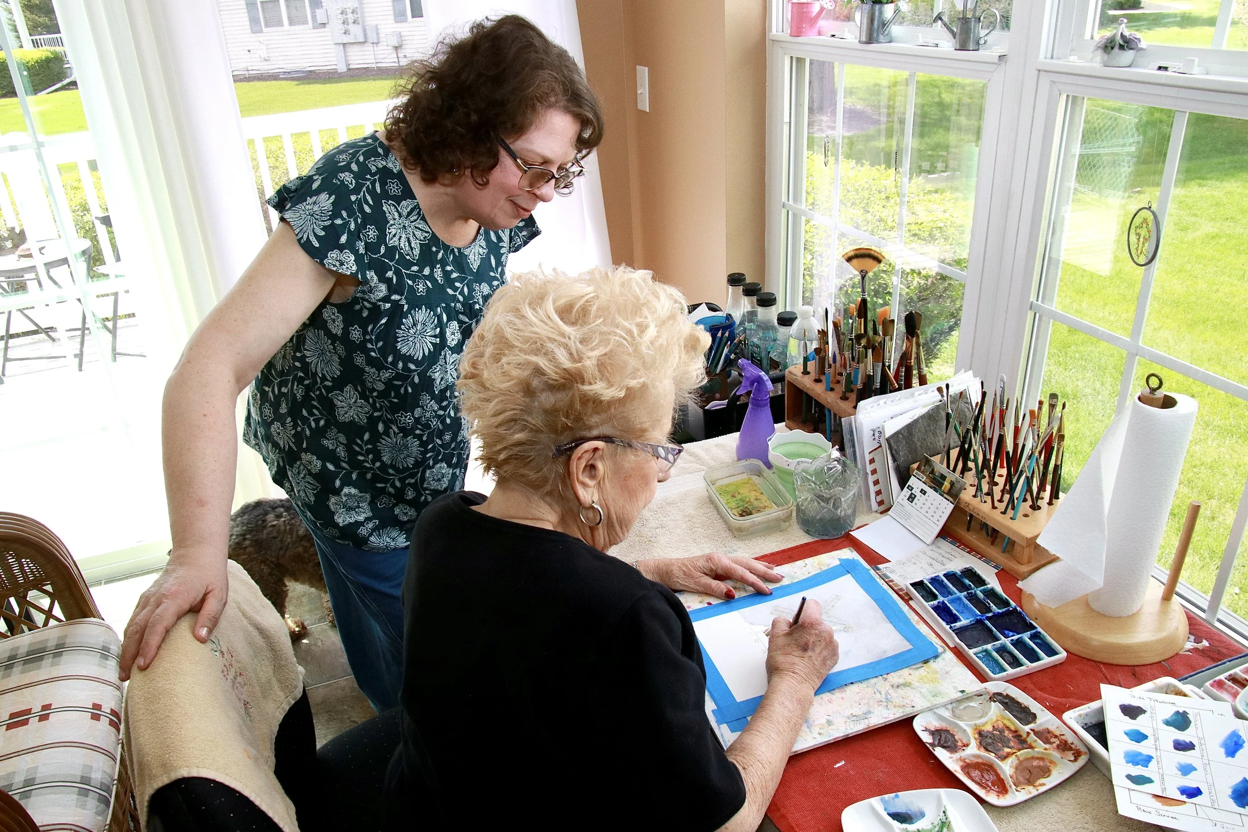Two women, one with gray hair and glasses, seated at a table painting watercolors, with art supplies nearby, while another woman with curly brown hair and glasses stands nearby, looking on in a sunlit room with large windows overlooking a green lawn.