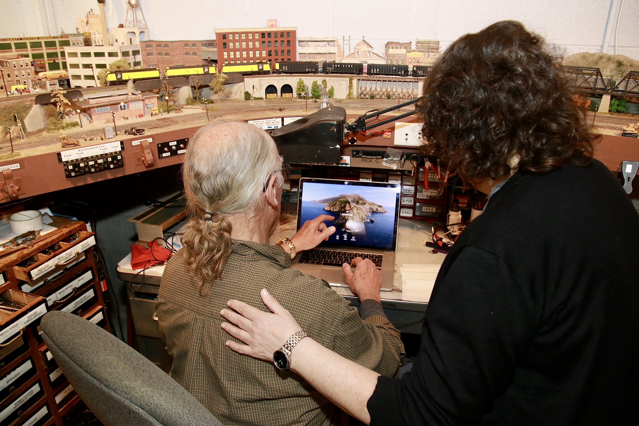 An older man and a women are discussing a model train layout in a room filled with model train scenery and train tracks, alongside a laptop and storage drawers.