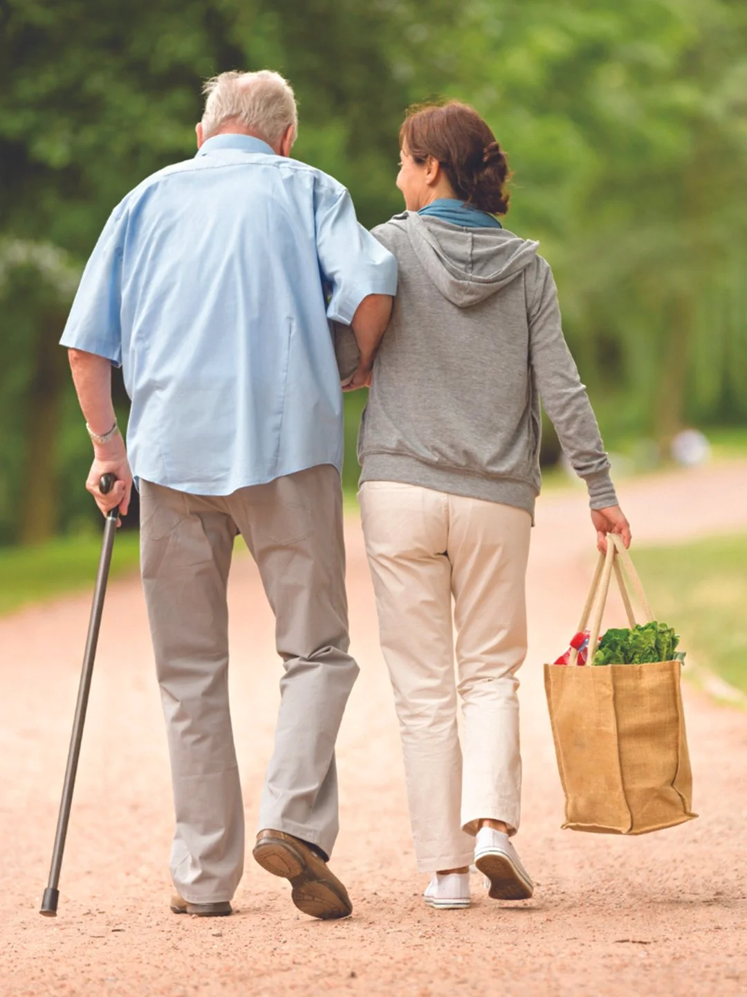 An elderly man with a cane and a woman walking together on a dirt path in a park, carrying a shopping bag filled with groceries.