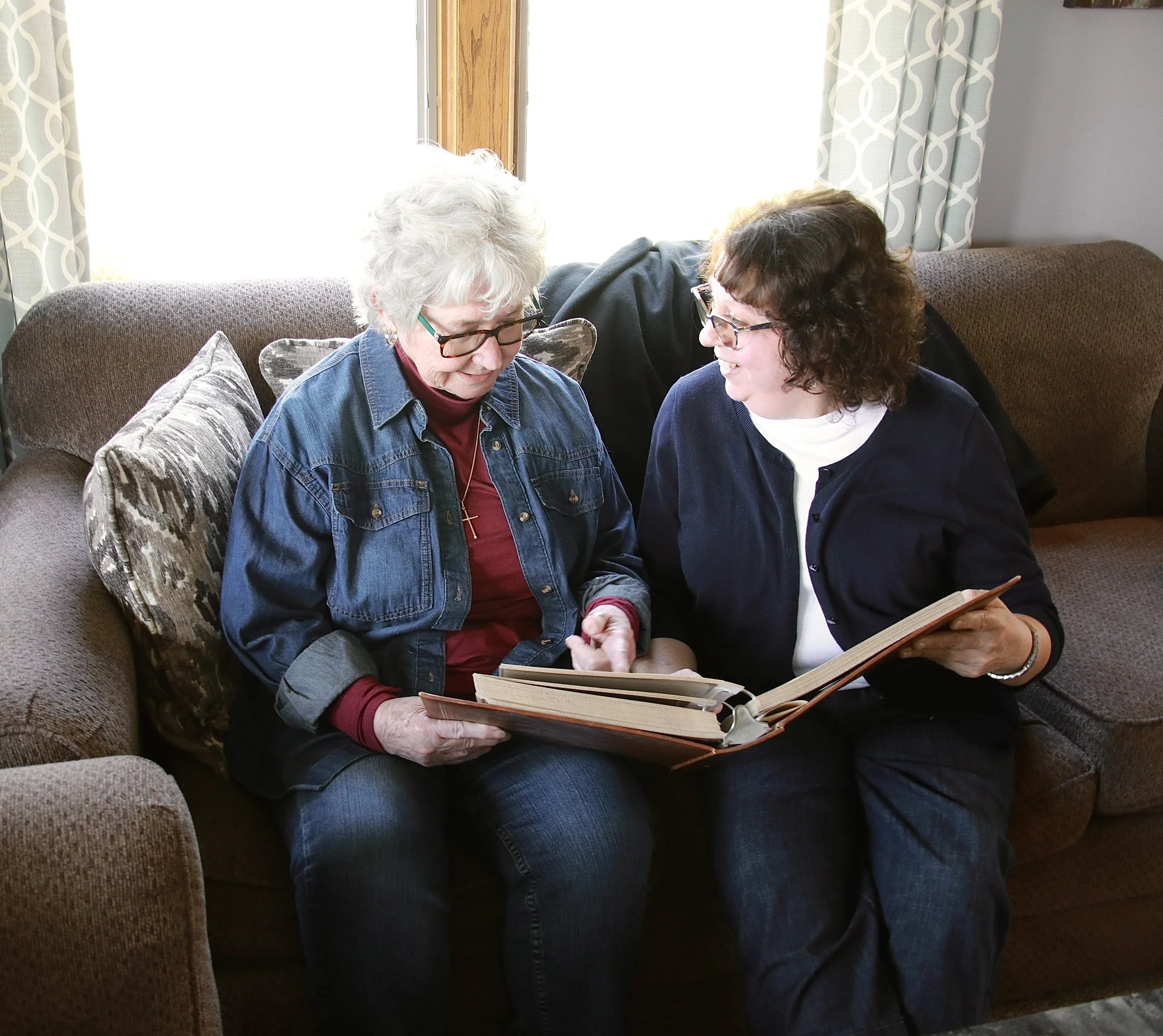 Two women sitting on a couch and looking at a photo album together in a cozy living room with sunlight streaming in.