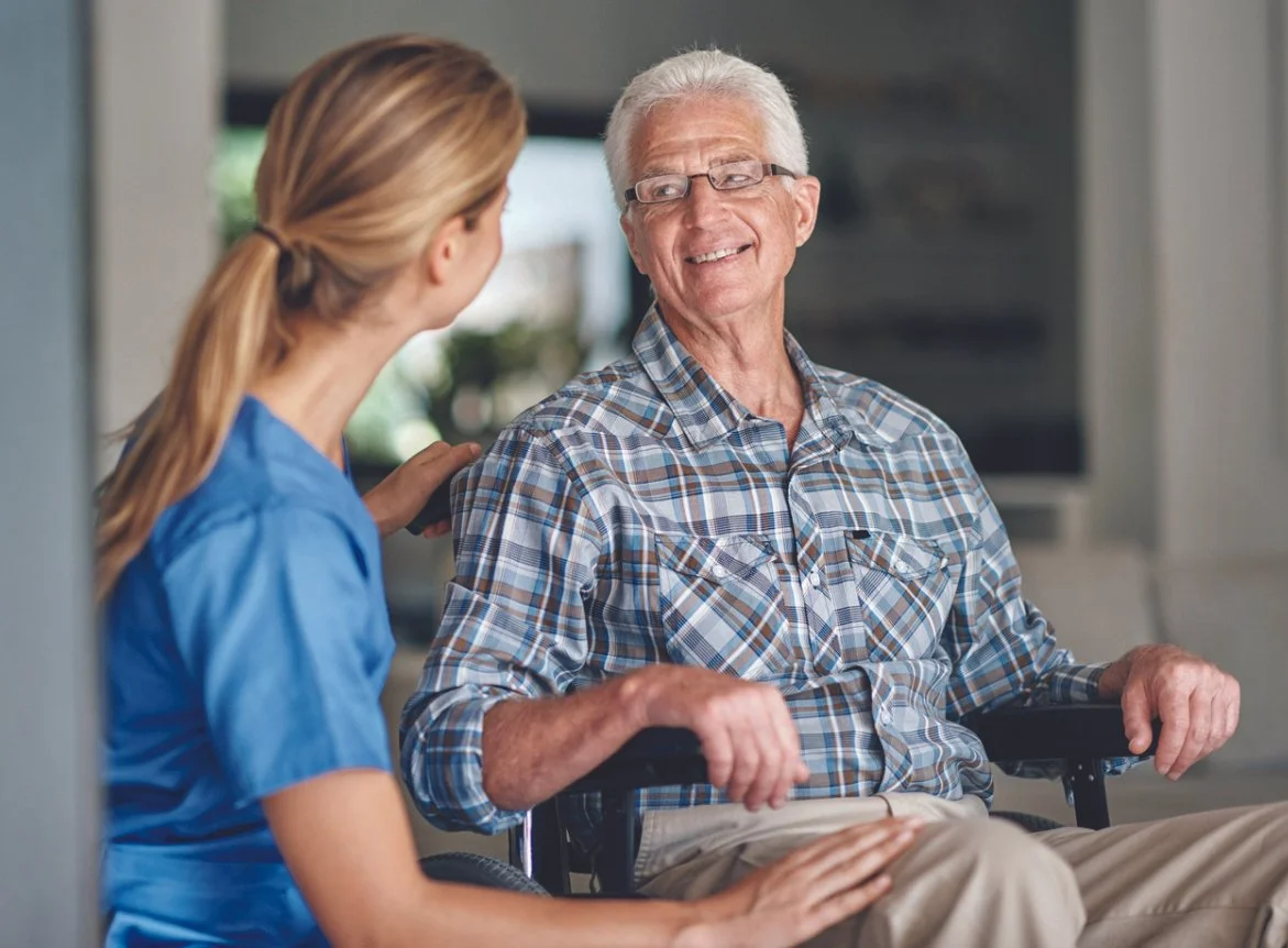 An elderly man in a wheelchair smiling and talking to a young woman in a blue shirt, in a bright, indoor setting.