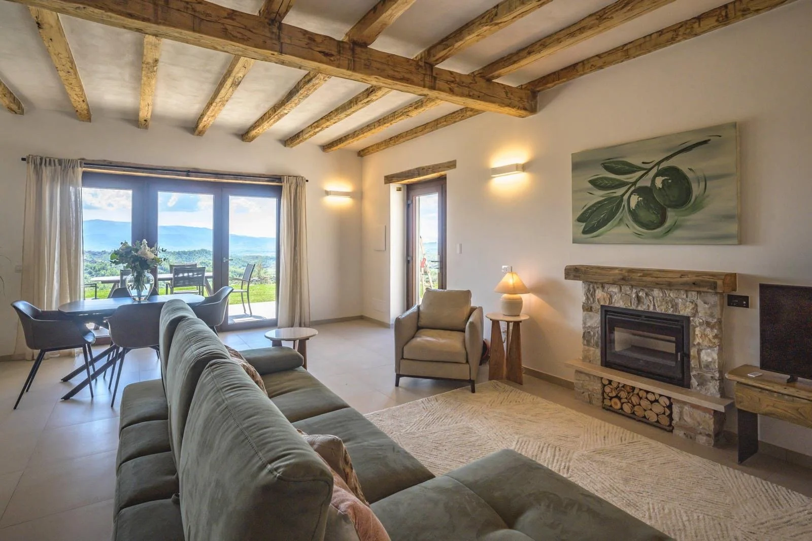 A living room with beige walls, a stone fireplace, a beige sofa, a beige armchair, a wooden side table with a lamp, and a painting of olives above the fireplace. Large glass sliding doors lead to a patio overlooking a green landscape and distant moun
