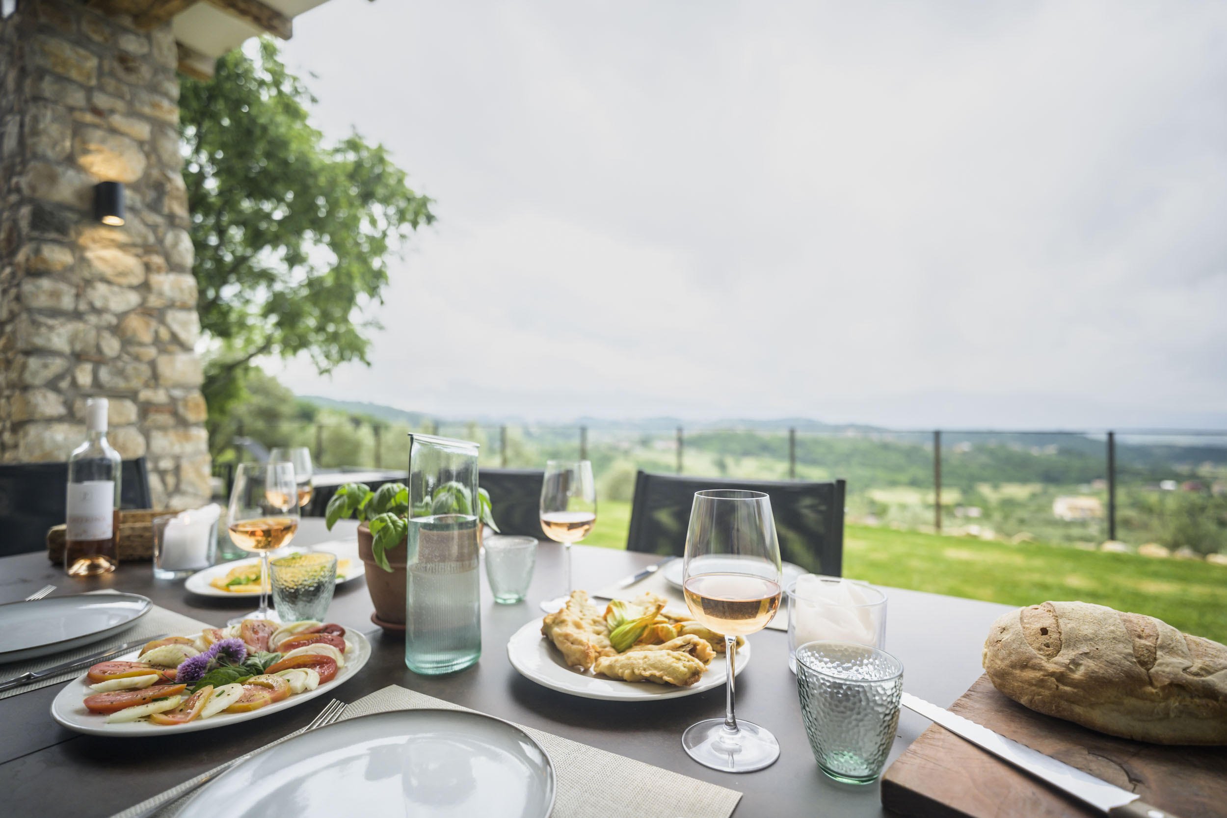Outdoor dining table set with wine glasses, plates of food including salad, baguette, pizza, and desserts, with a scenic view of green hills and cloudy sky in the background.