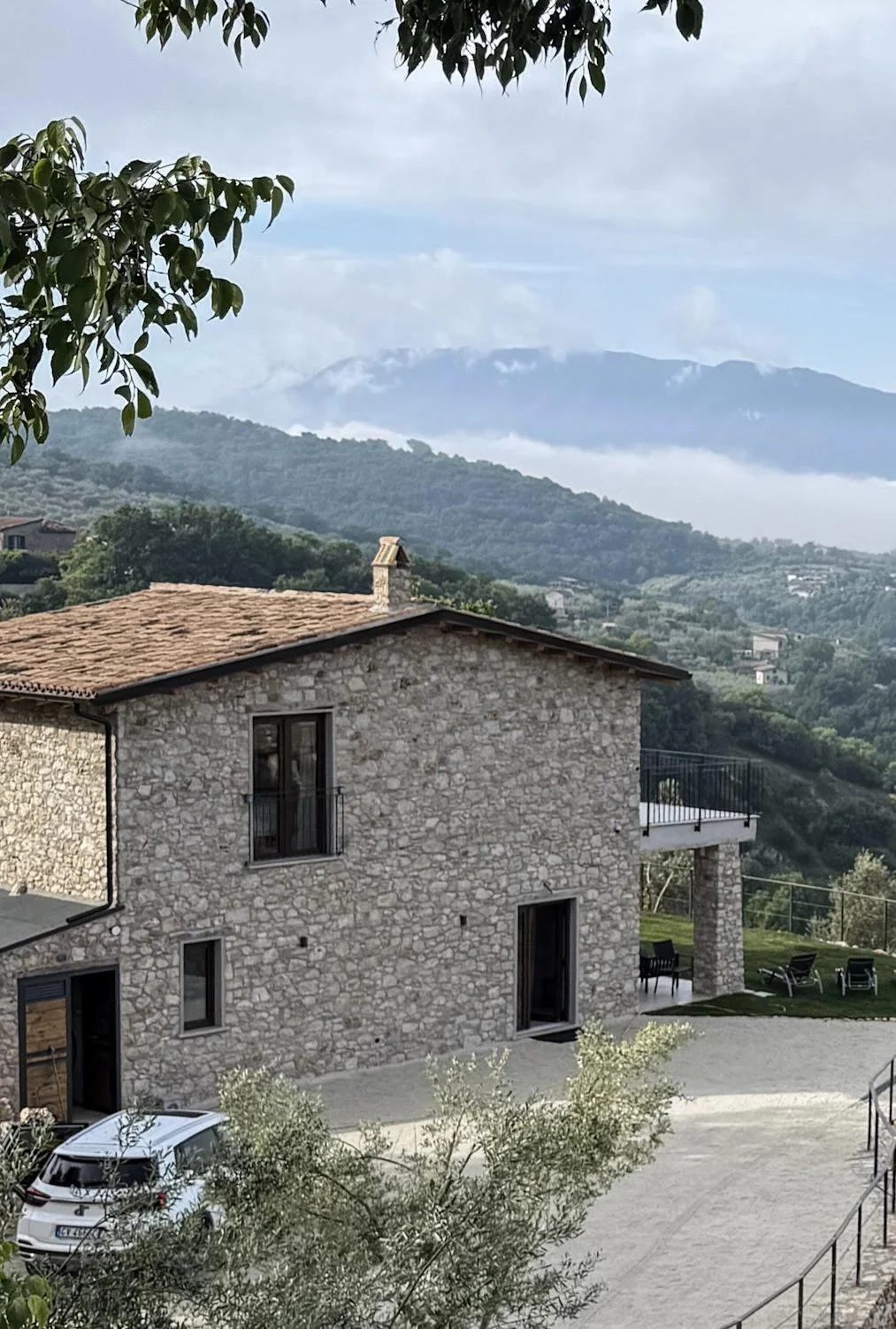 A stone house with a tiled roof surrounded by greenery and hills, with mountains in the background and a car parked nearby.