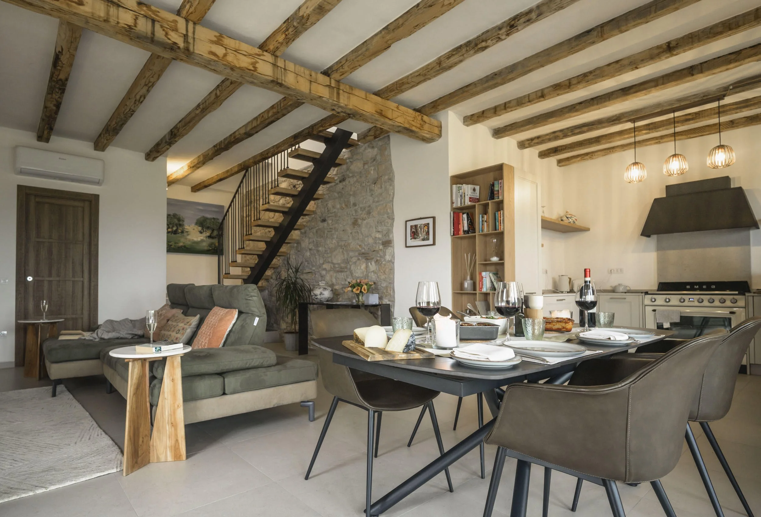 Living and dining area with exposed wooden beams on the ceiling, a stone wall with art, a grey sofa with pillows, a table with glasses, and a dining table set with food and wine. There is a kitchen visible in the background.