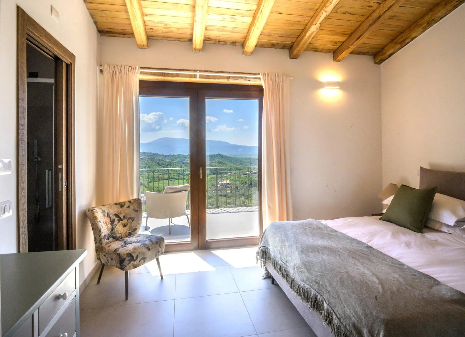 Hotel room with a sliding glass door opening to a balcony showing a scenic mountainous landscape and blue sky with clouds, a bed with pillows, a floral armchair, and a chair on the balcony.
