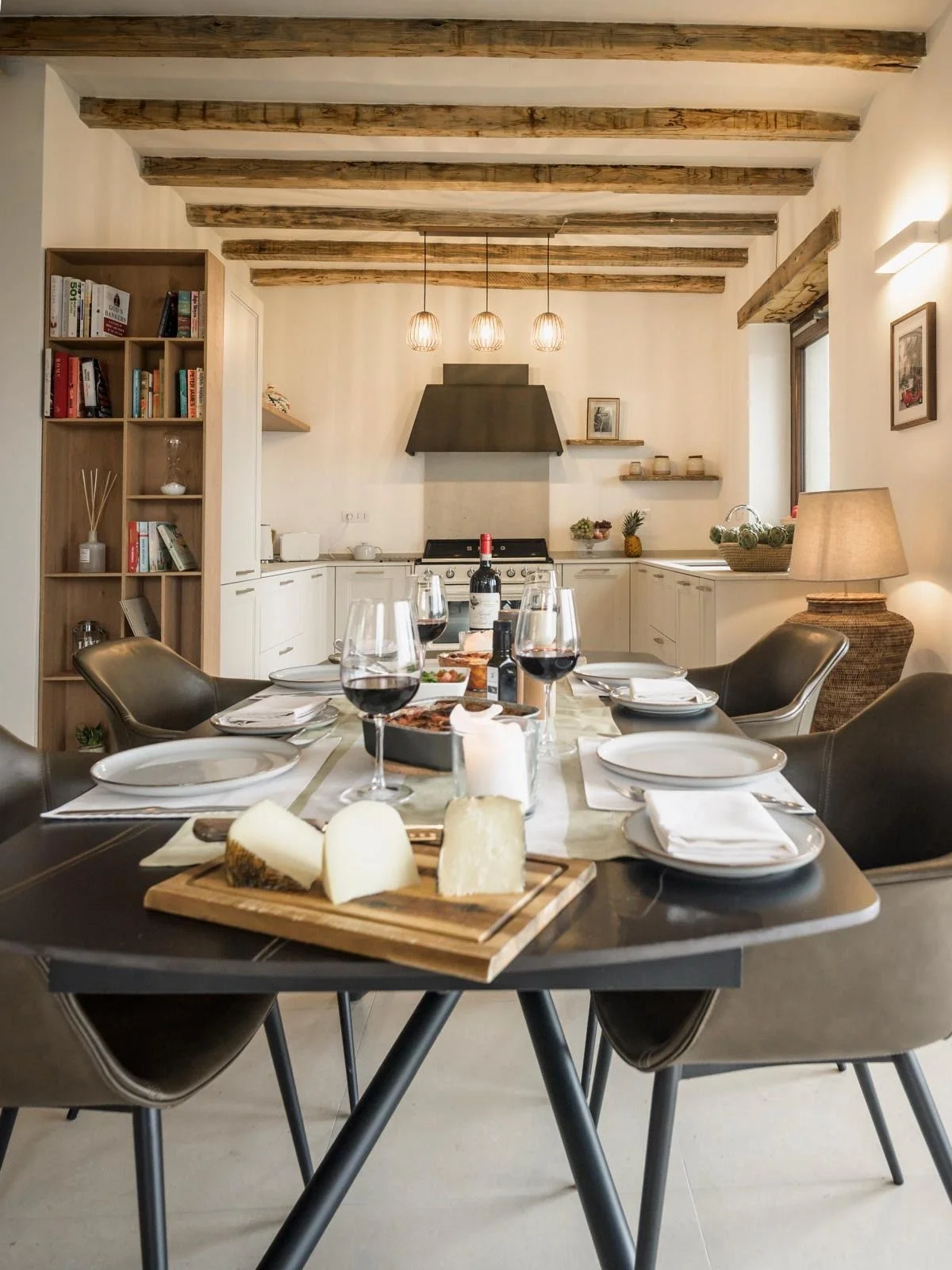 A dining table set for a meal with wine glasses, cheese, and plates in a cozy, rustic kitchen with exposed wooden beams and modern decor.